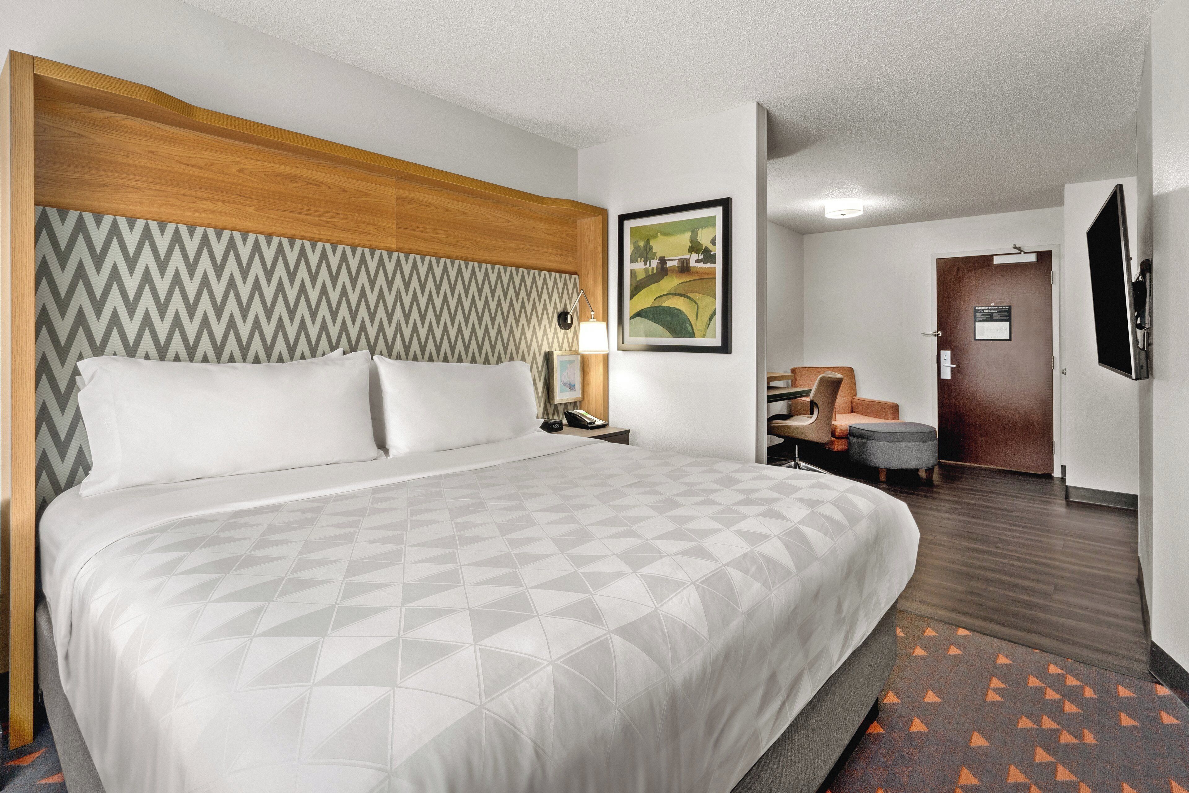 A hotel room with a large bed, a wooden headboard, a desk with a chair, a television, and a painting on the wall.