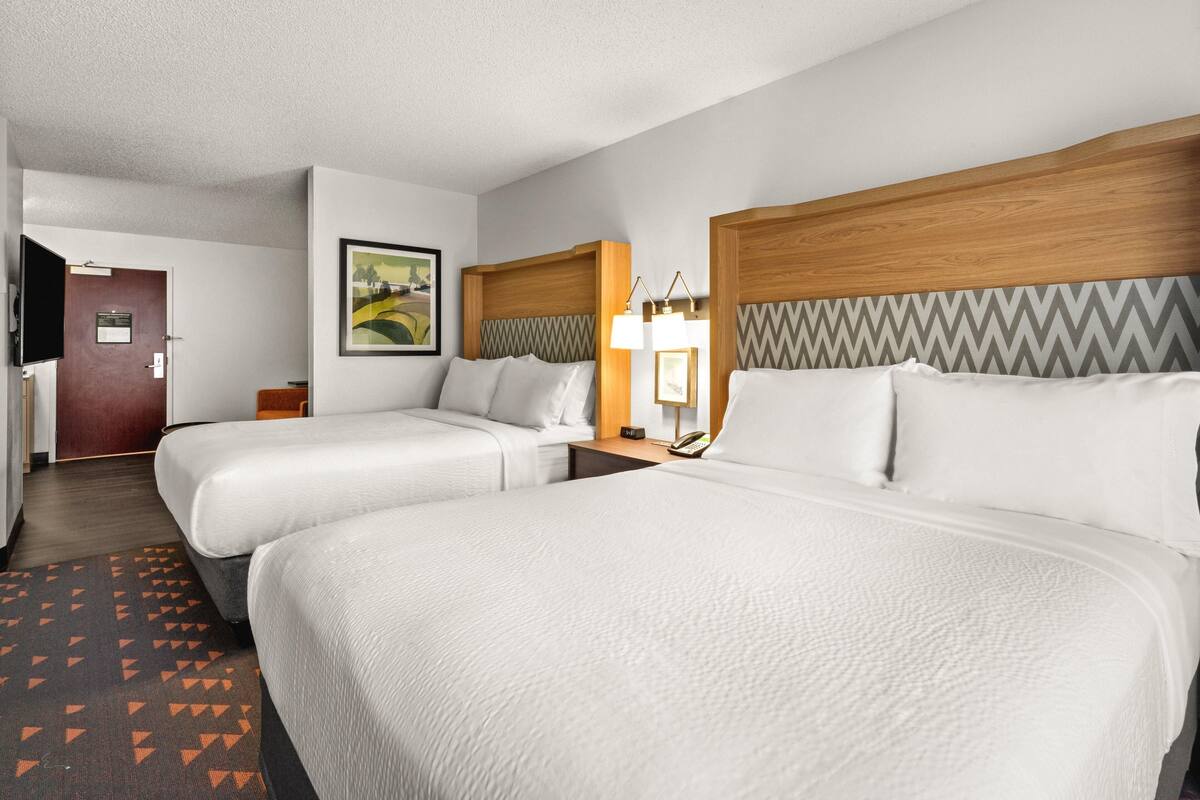 A hotel room with two beds, a wooden headboard, a nightstand with a lamp, a framed picture on the wall, and a door leading to another room.