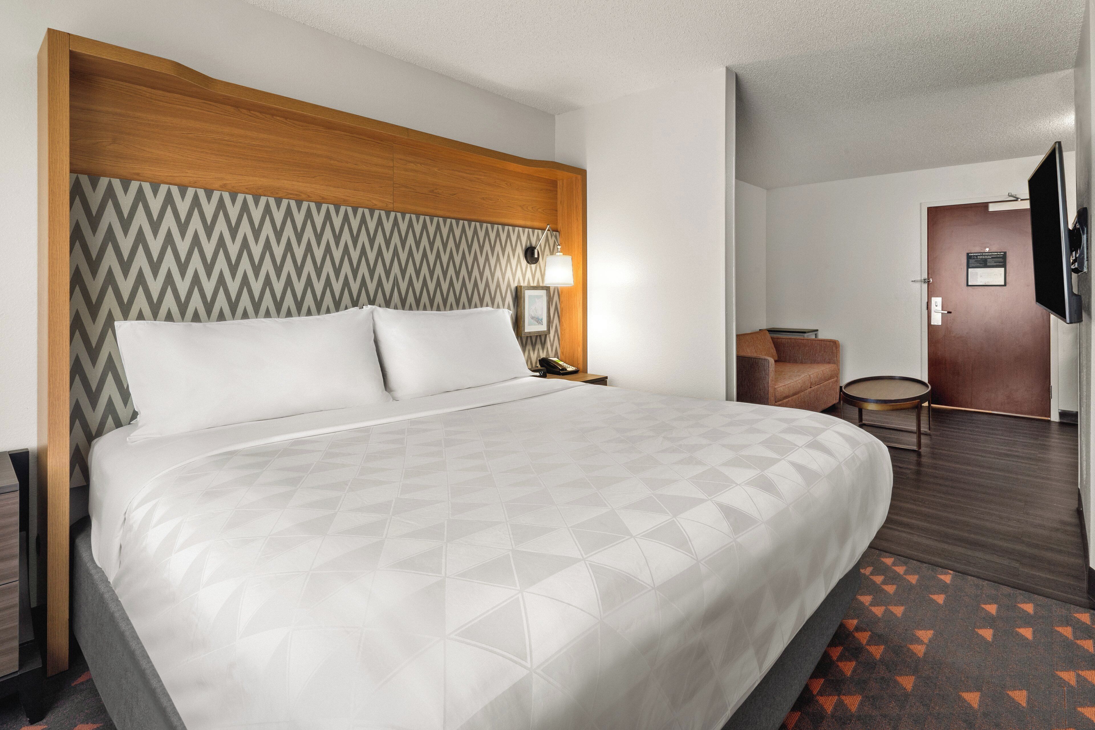 A hotel room with a large bed, a wooden headboard, a bedside table with a lamp, a chair, and a television mounted on the wall.