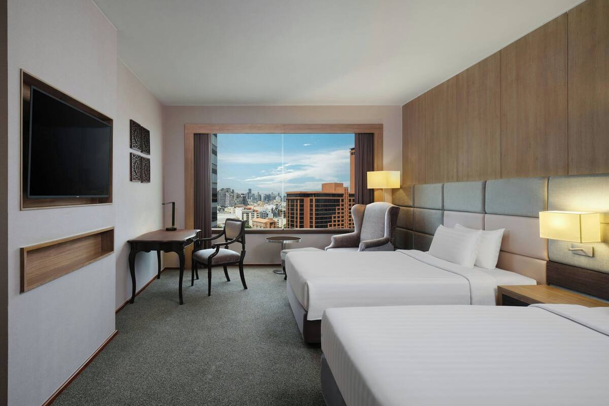A hotel room with two beds, a desk, a chair, a TV, and a window with a city view.
