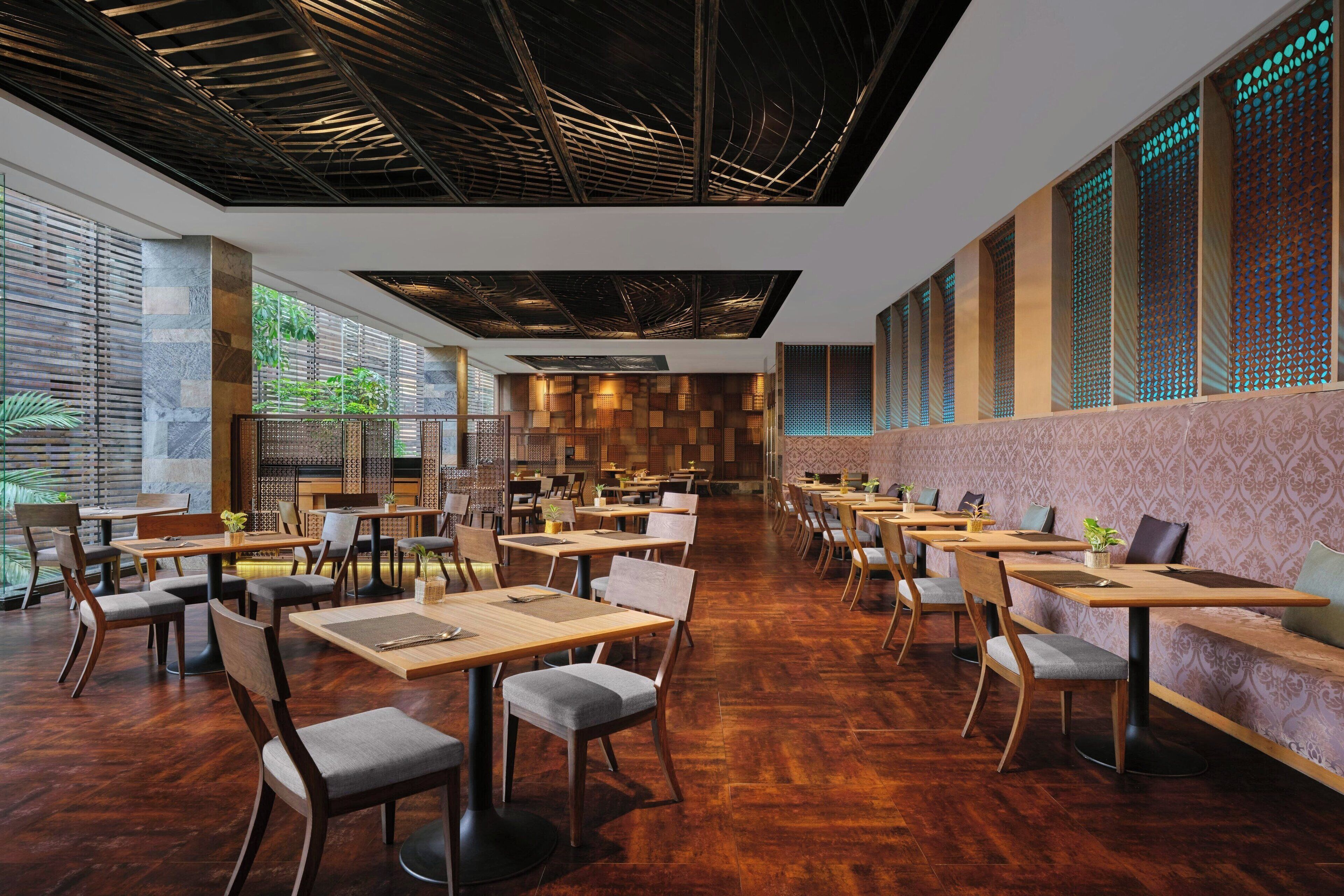A spacious restaurant with wooden tables and chairs, a high ceiling with exposed beams, and large windows allowing natural light.