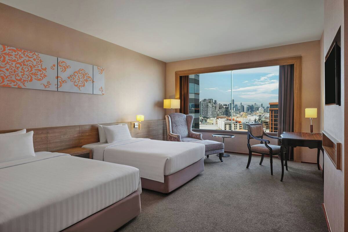 A hotel room with two beds, a desk, a chair, and a view of a cityscape through large windows.