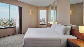 A hotel room with a large bed, a nightstand, a mirror, and a view of the cityscape through large windows.
