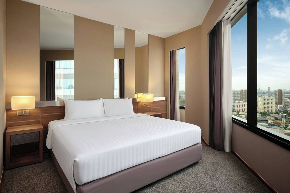 A modern hotel room with a large bed, two bedside tables with lamps, a large mirror, and a city view through a large window.