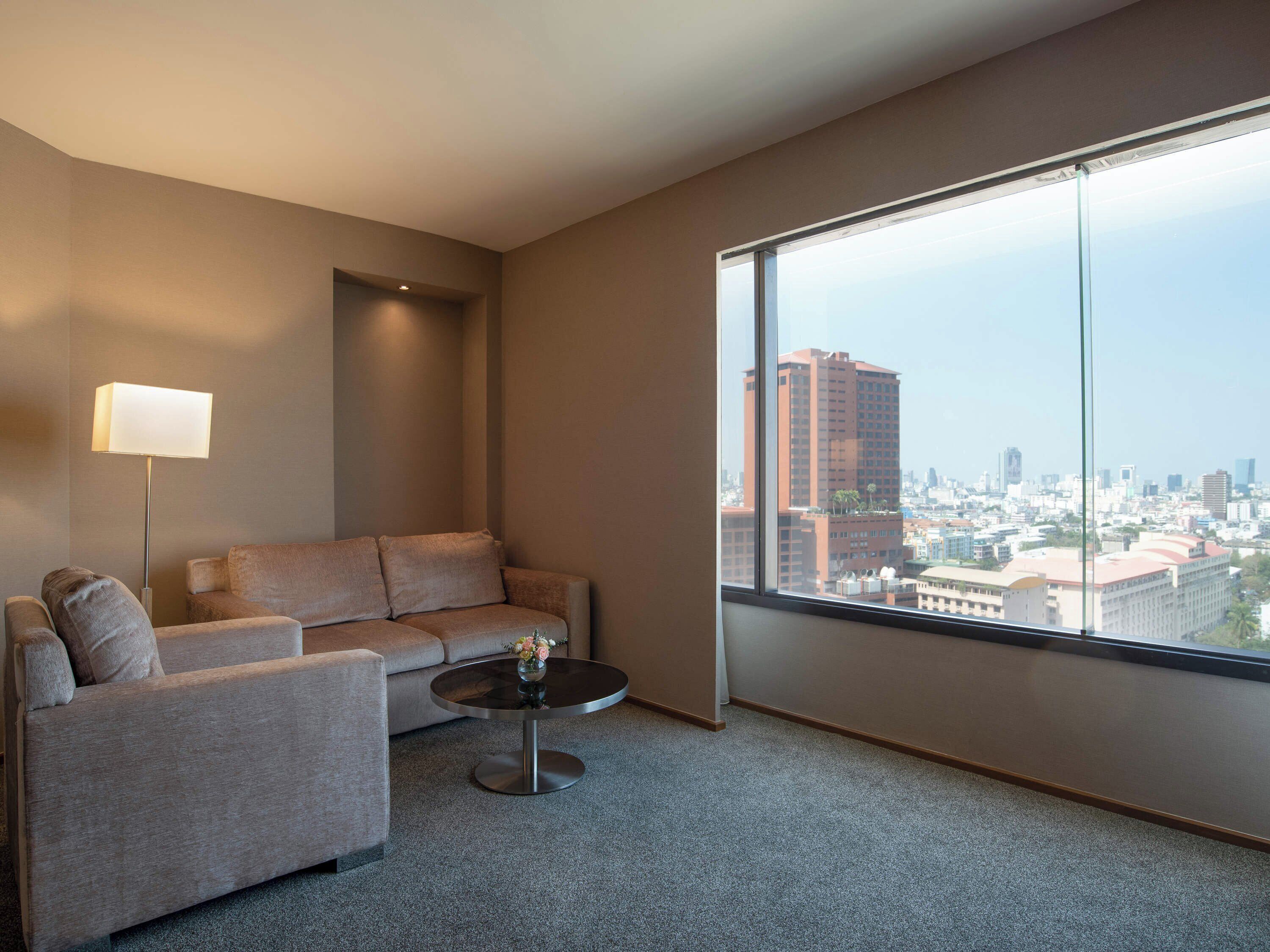 Habitació d'hotel moderna amb una finestra gran que ofereix vista a la ciutat, un sofà, una taula rodona petita i una làmpada de peu.