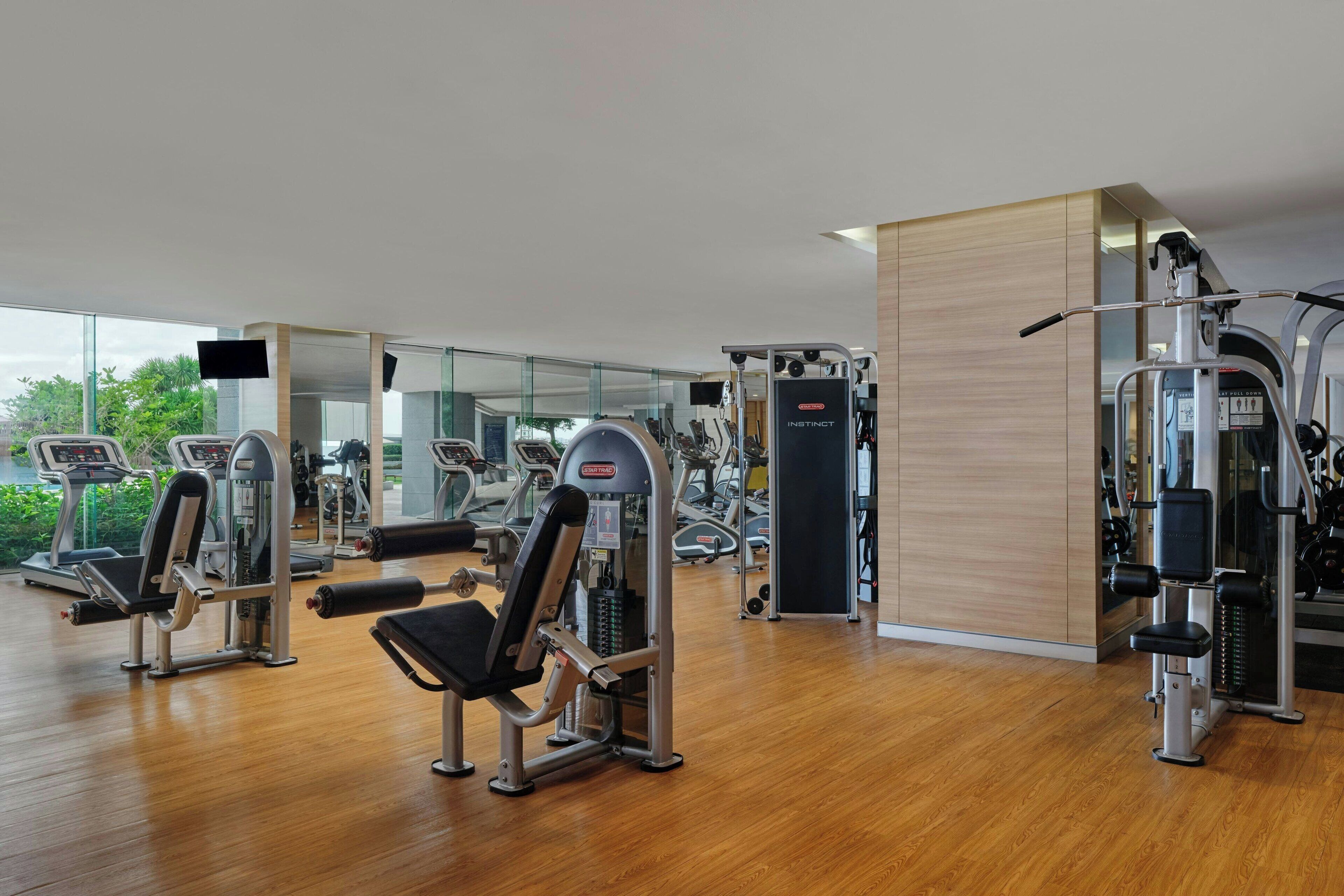 A modern gym with various exercise machines, large windows, and a wooden floor.