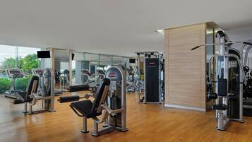 A modern gym with various exercise machines, large windows, and a wooden floor.
