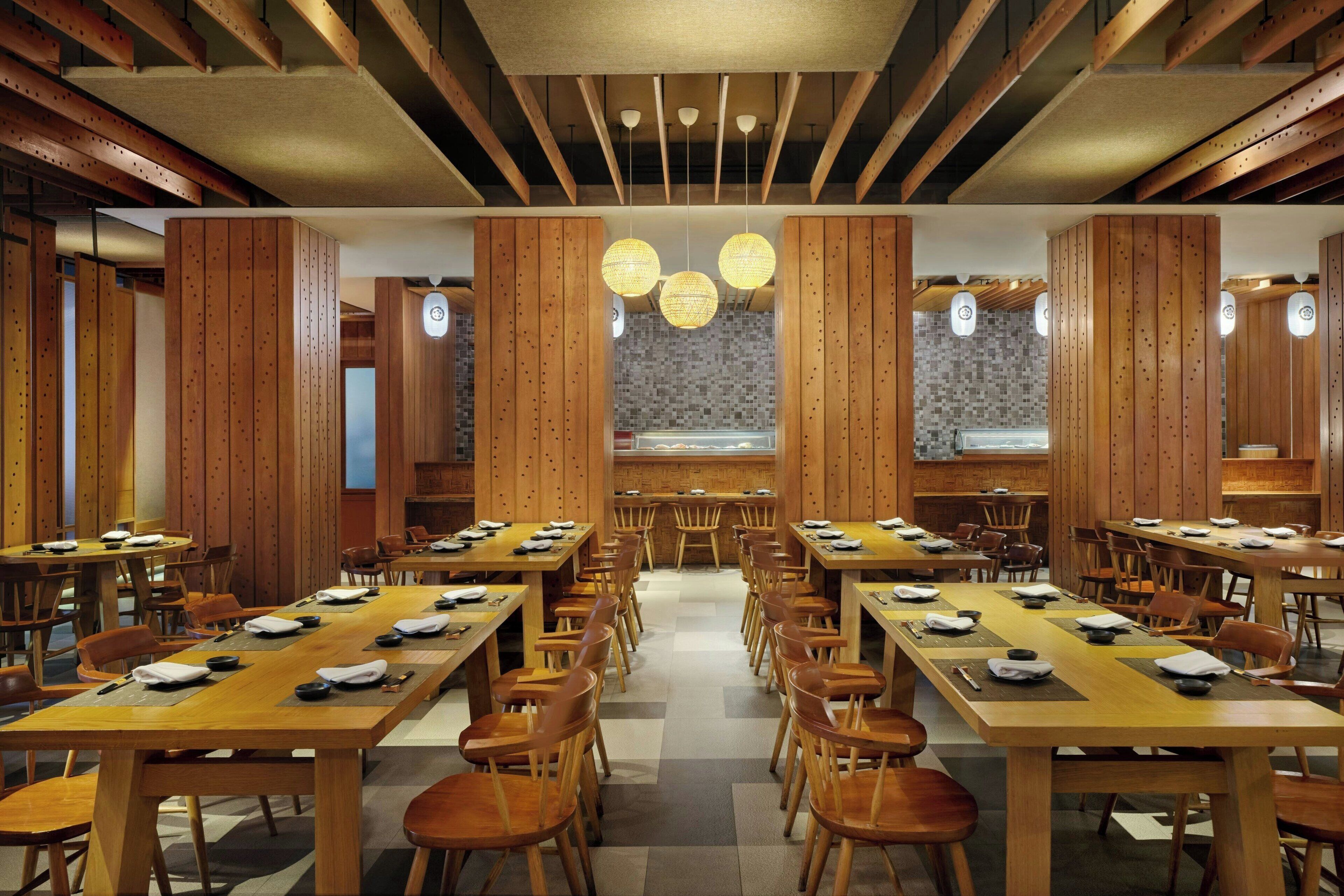 A spacious dining area with long wooden tables and chairs, centered around a patterned wall and a view of the city.