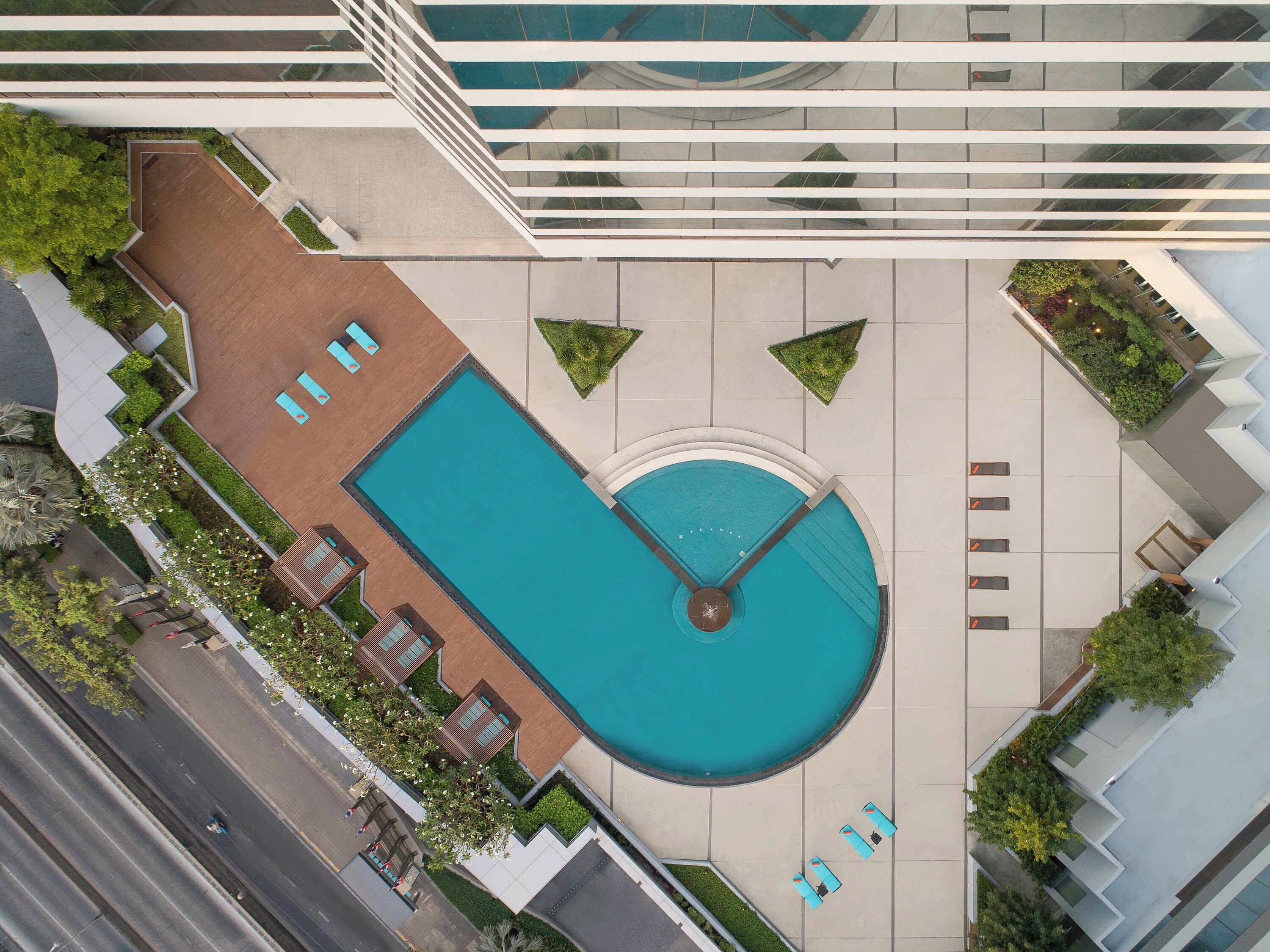 An aerial view of a modern building with a large rectangular swimming pool, surrounded by lounge chairs and small landscaped areas.