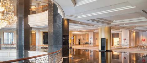 Un amplio vestíbulo de hotel con una gran araña, suelos de mármol pulido y un balcón curvo con barandillas ornamentales.