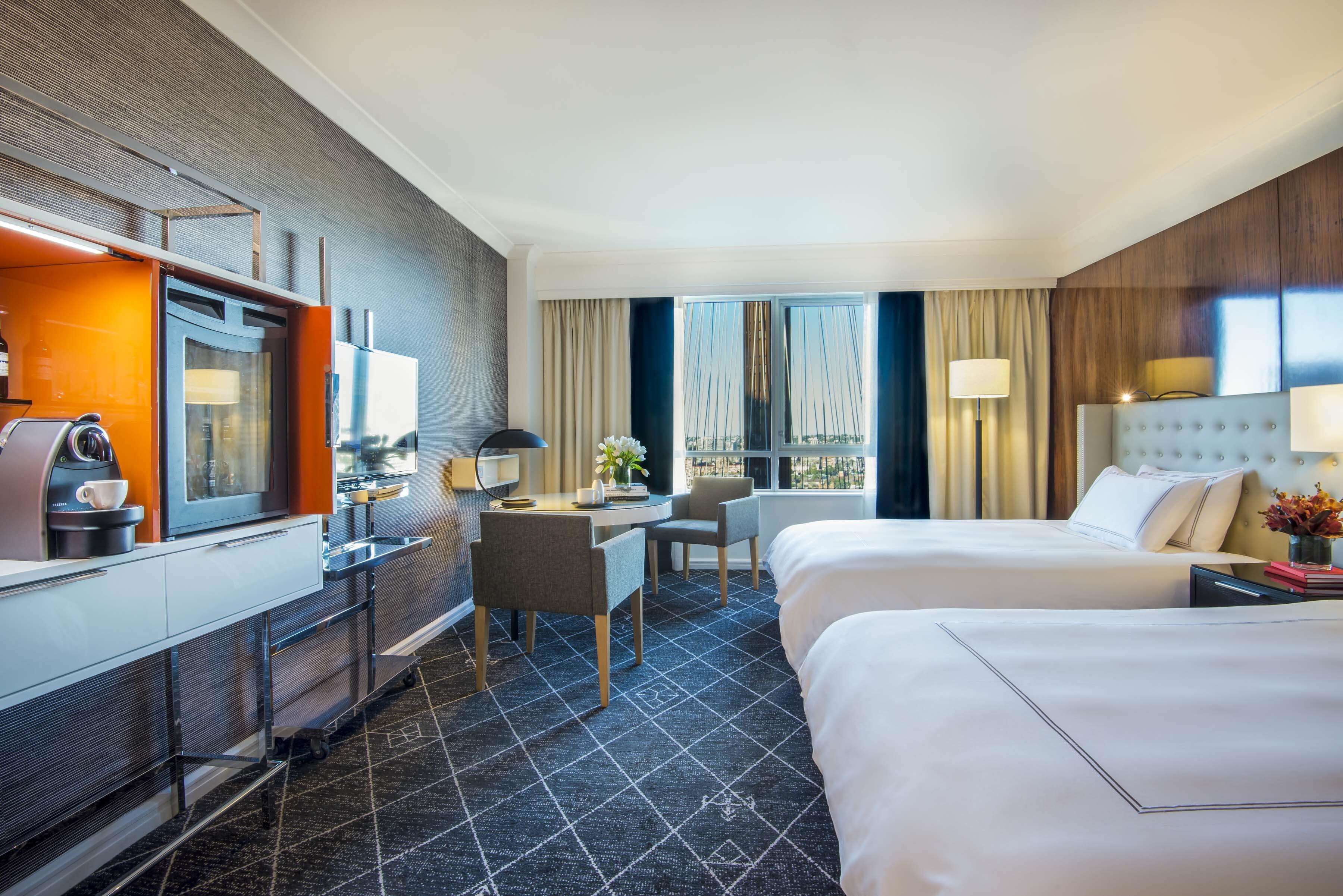 A hotel room with two beds, a desk, a chair, a coffee machine, and a view of the city through large windows.