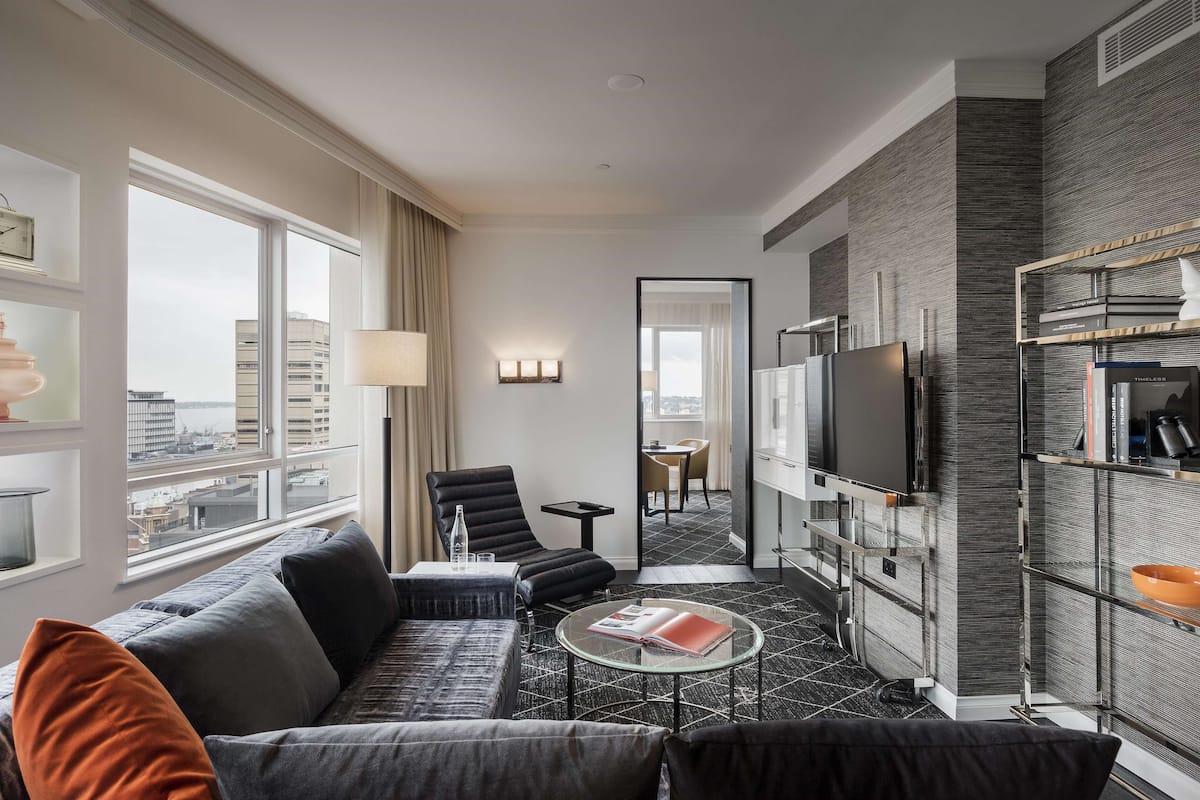 Una habitación de hotel moderna con sofá, sillón, mesa de centro, televisor y estantería.