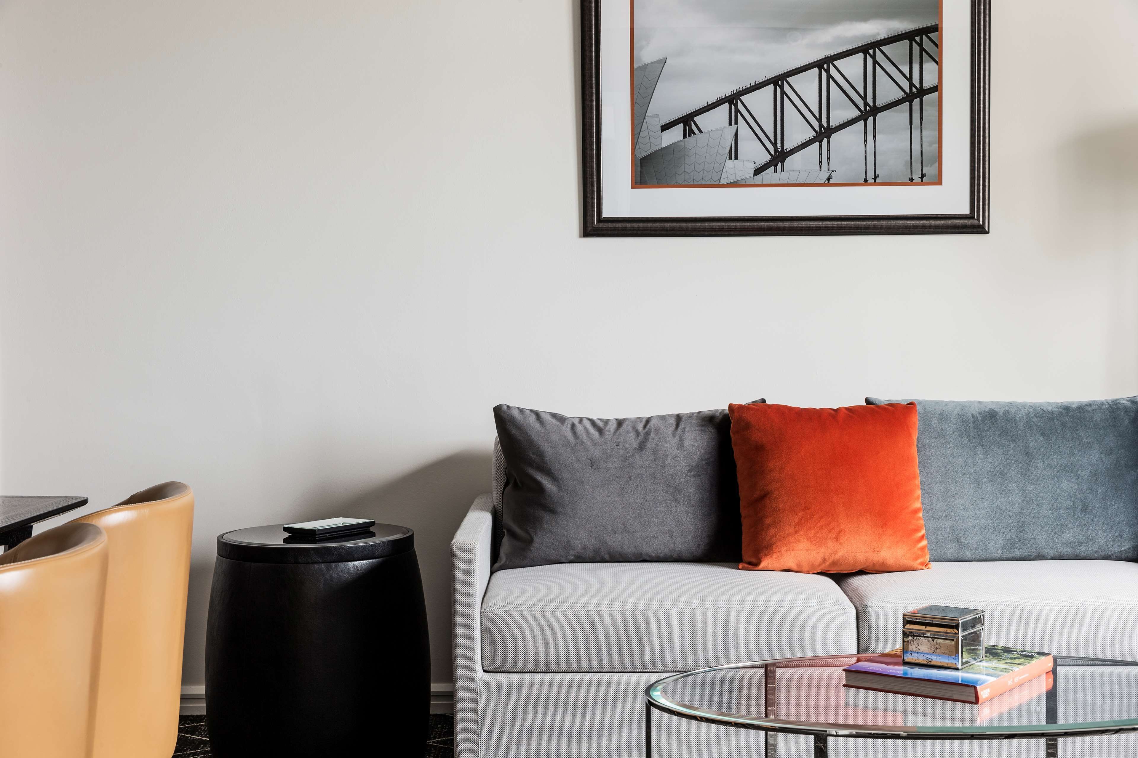 A modern living room with a sofa, two pillows, a glass coffee table with books, a black side table, and a framed picture on the wall.