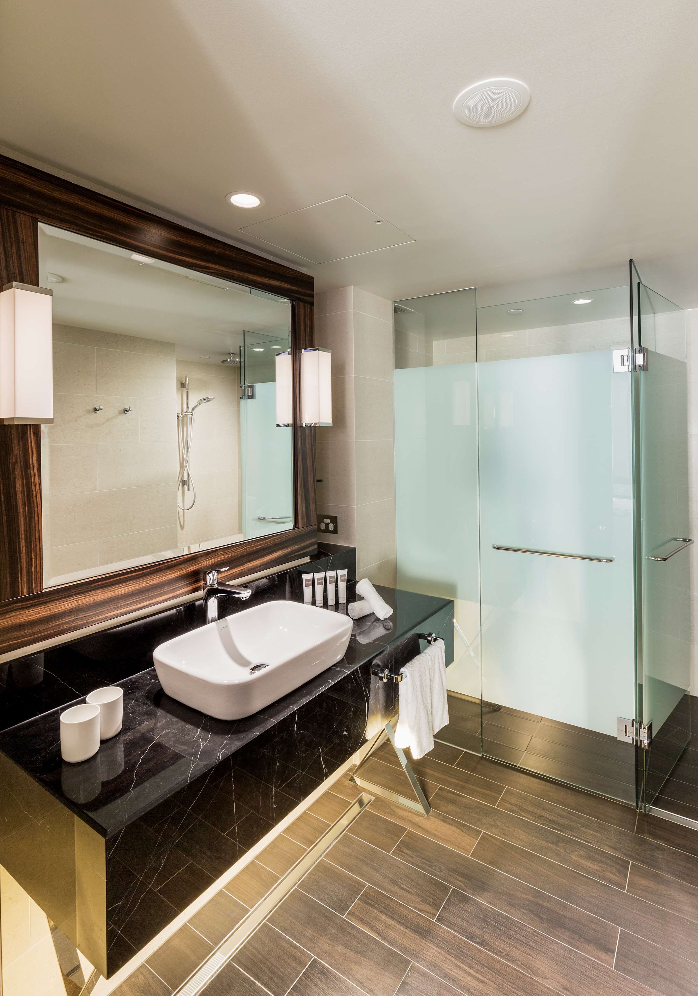 A modern bathroom with a large mirror, a white sink, a glass shower enclosure, and dark wood flooring.