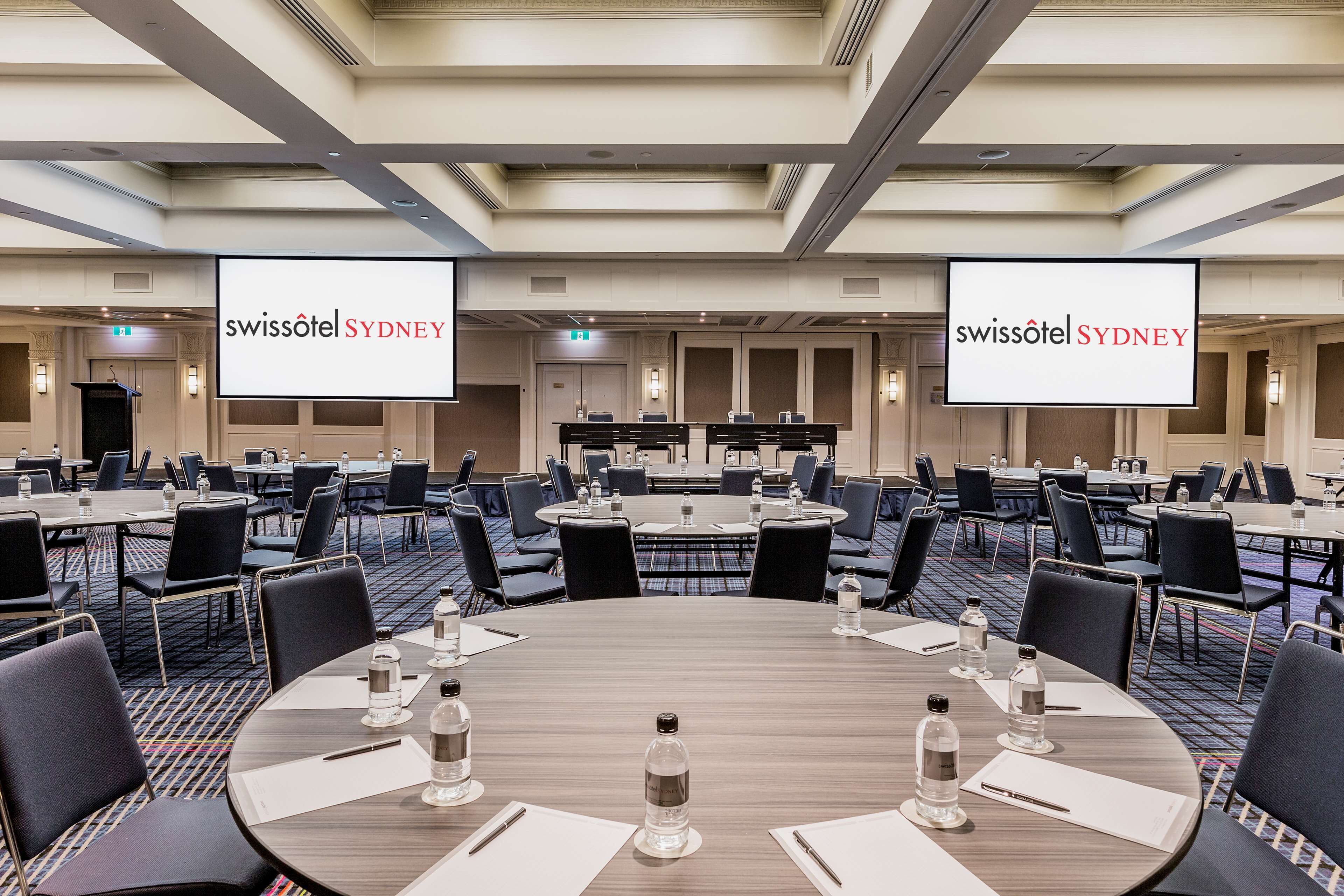 Un salón de conferencias con varias mesas, sillas y una pantalla grande que muestra el nombre del hotel.
