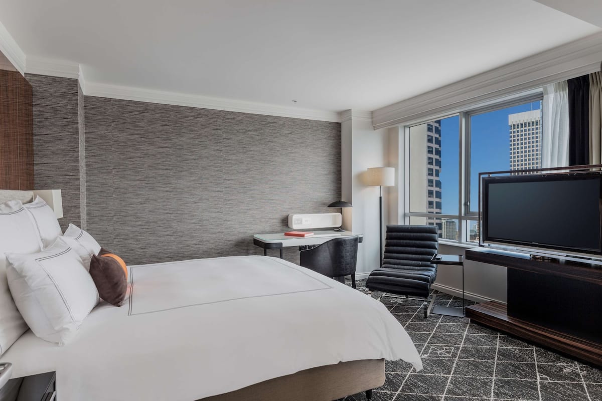 Habitación de hotel con una cama grande, un escritorio con lámpara, una silla, un televisor y vistas de la ciudad por la ventana.