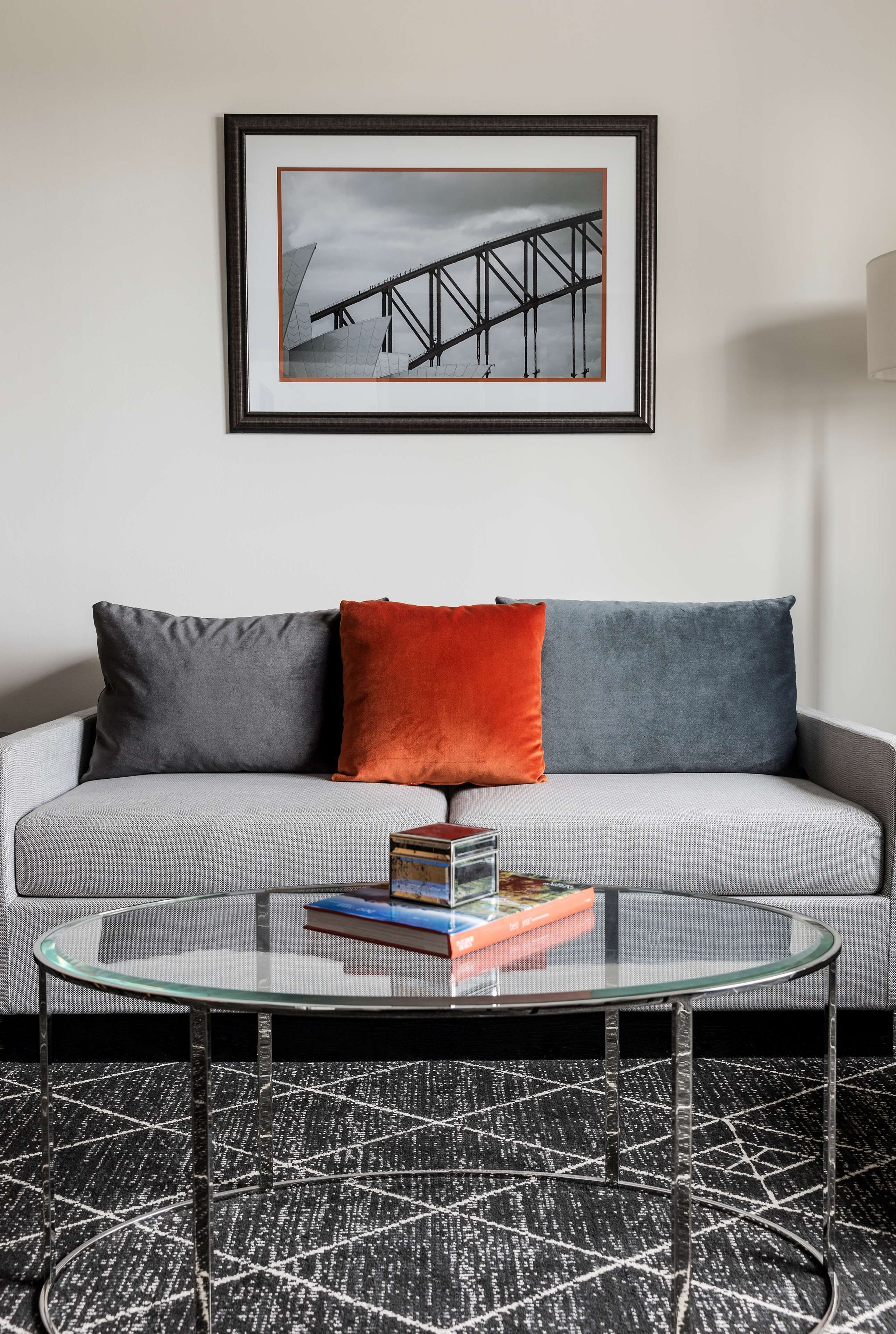 A modern living room with a grey sofa, two cushions, a glass coffee table, and a framed picture on the wall.