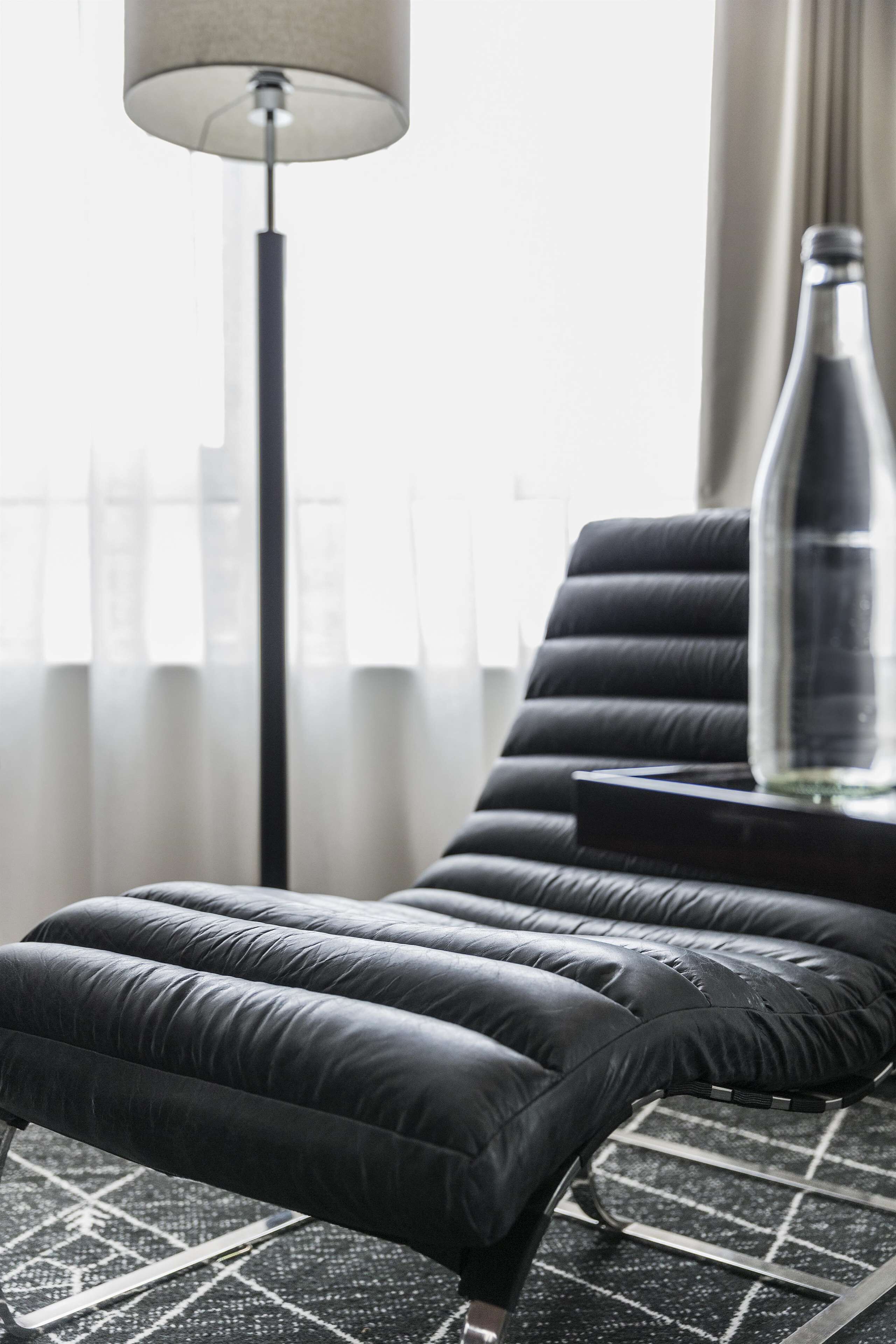 A modern lounge chair with a black leather seat and backrest, positioned on a patterned rug. A floor lamp with a cylindrical shade is visible in the background, and a clear glass bottle rests on a small table beside the chair.