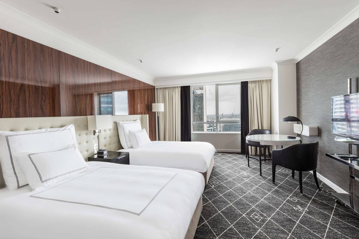 Habitación de hotel con dos camas, un escritorio, una silla, un televisor y un ventanal con vistas a la ciudad.