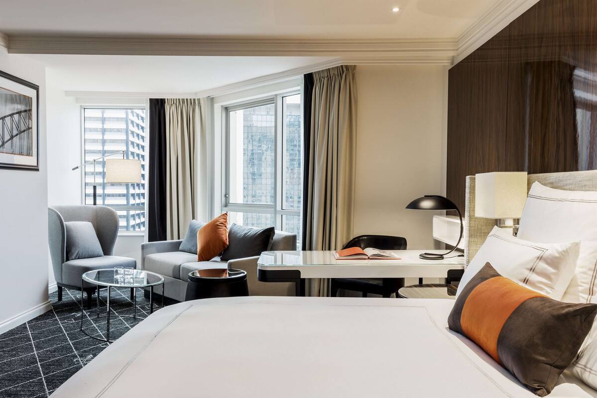 Una habitación de hotel con cama, dos sillones, una mesita, un escritorio y una ventana con cortinas.