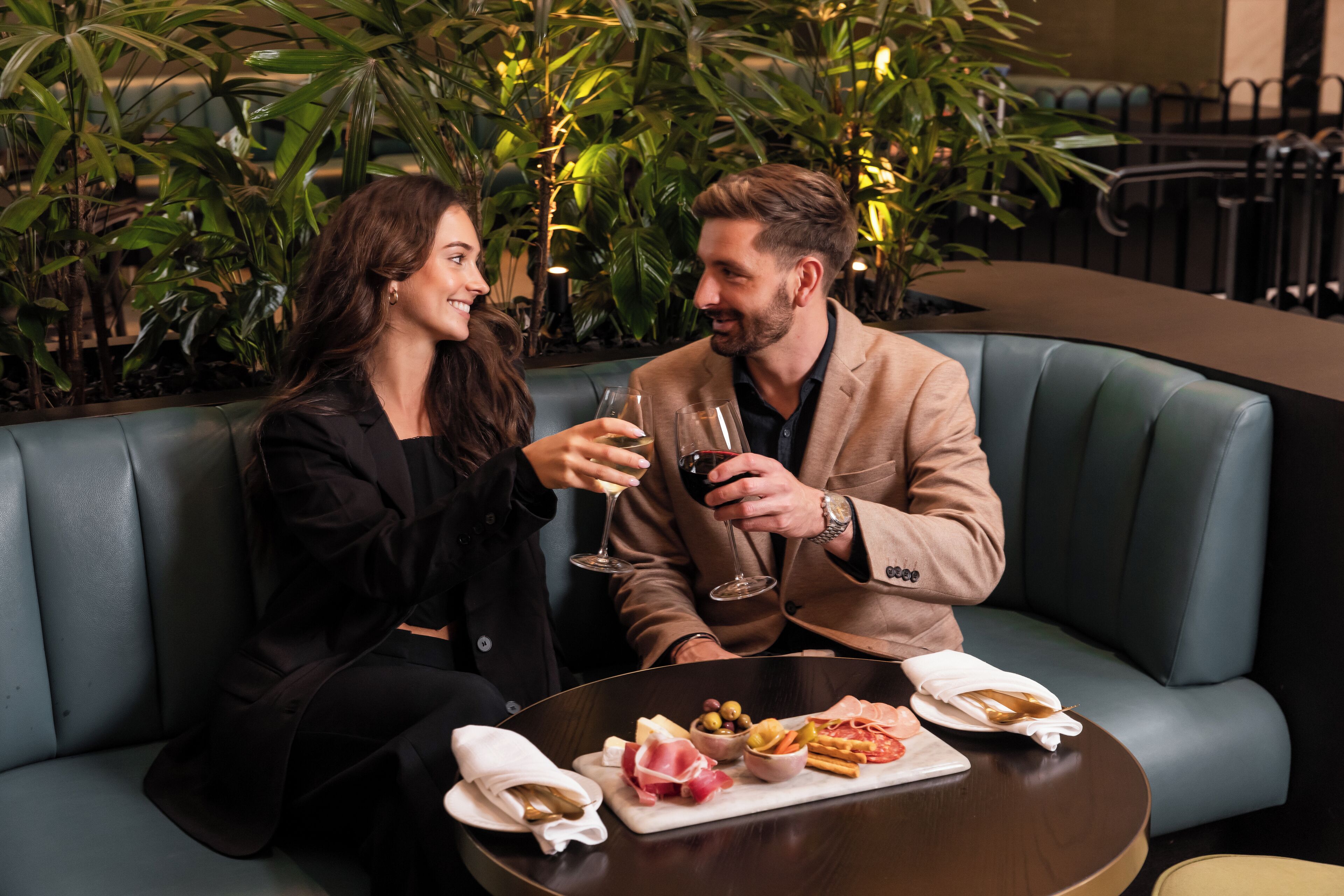 Two people seated on a curved sofa with a table in front of them, holding wine glasses.