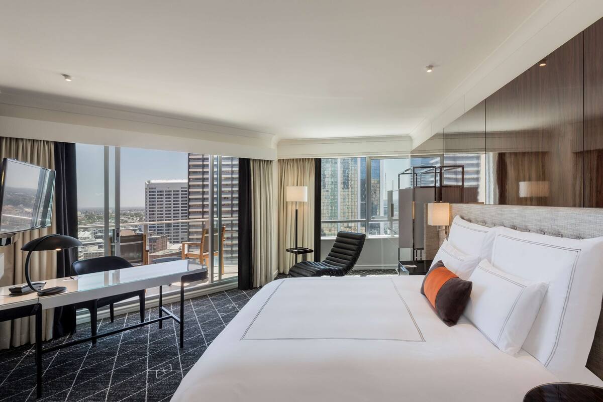 Habitación de hotel con una cama grande, un escritorio, una silla y un televisor. La habitación tiene ventanas amplias con vista a la ciudad.