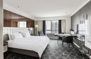 A modern hotel room with a large bed, a desk, a chair, a TV, and a view of the city. - Swissotel Sydney (Sydney)