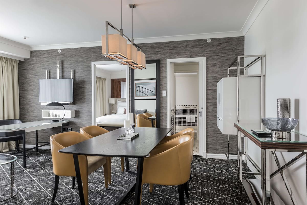 Habitación de hotel moderna con zona de comedor, cama, televisor y baño visible a través de una puerta abierta.