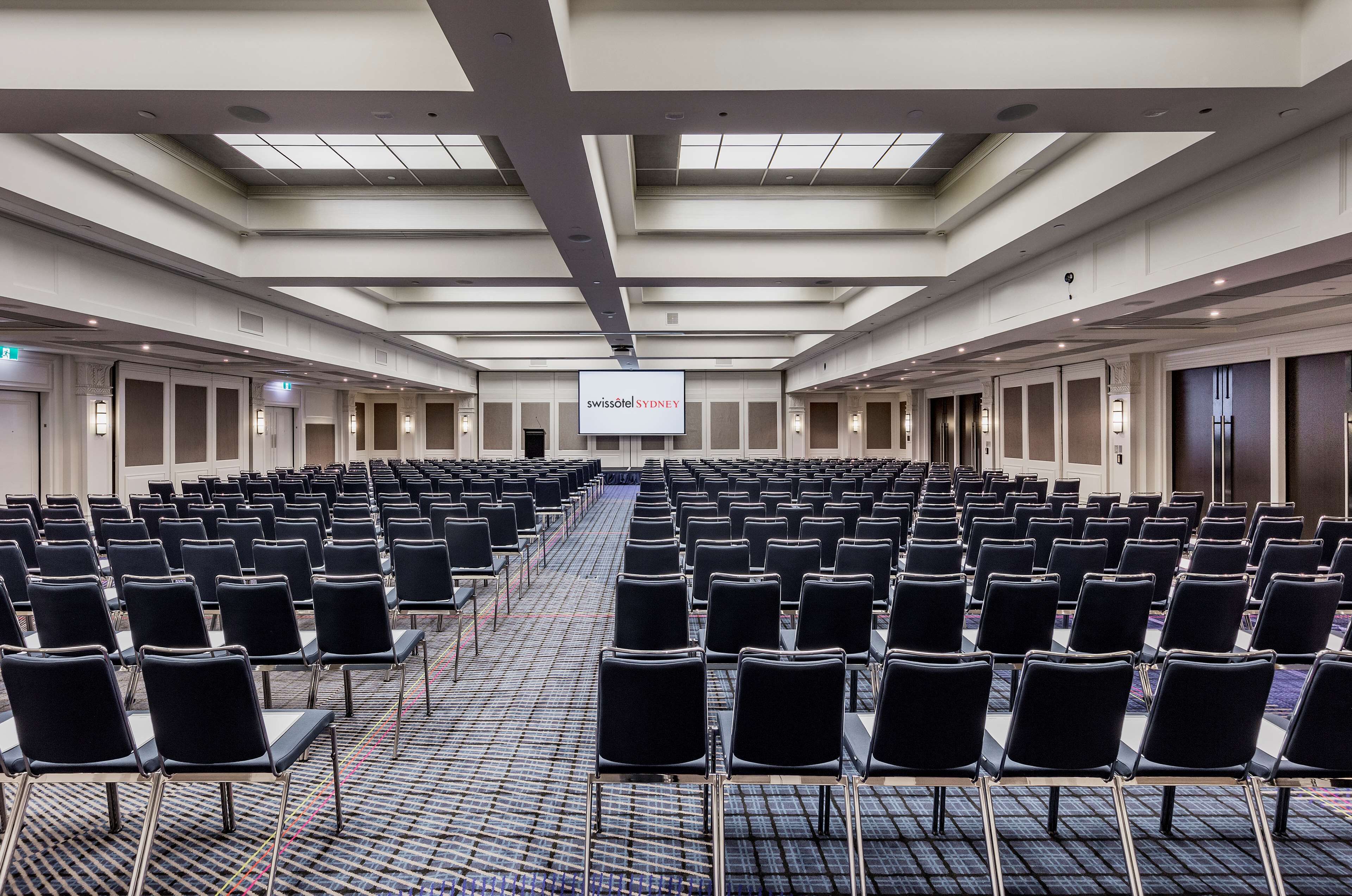Une salle de conférence avec des rangées de chaises noires, un grand écran affichant le nom « swissotel SYDNEY », et un plafond haut doté de lucarnes.