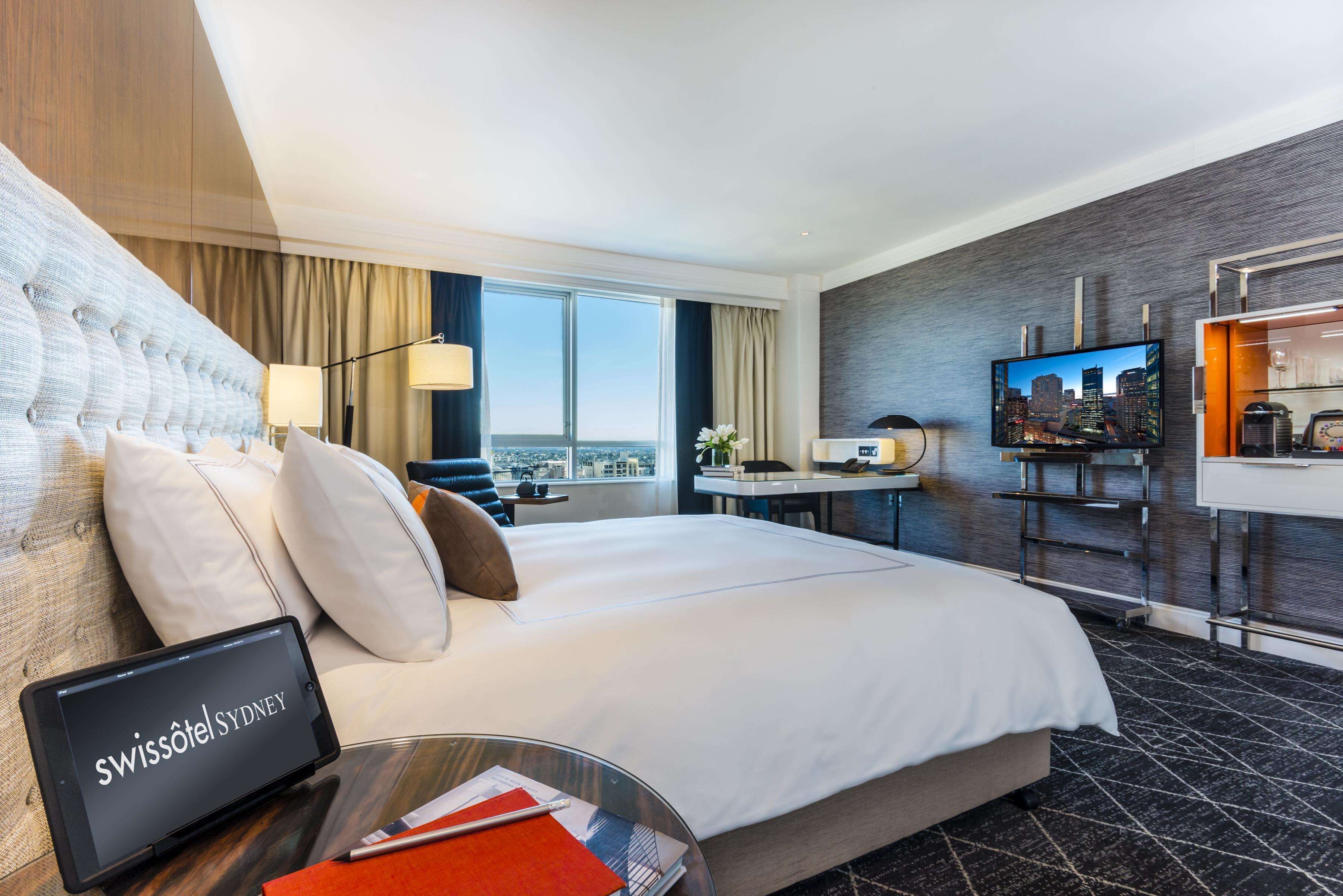 Ein modernes Hotelzimmer mit einem großen Bett, einem Schreibtisch, einem Fernseher und einem Laptop, der "Swissôtel Sydney" anzeigt.