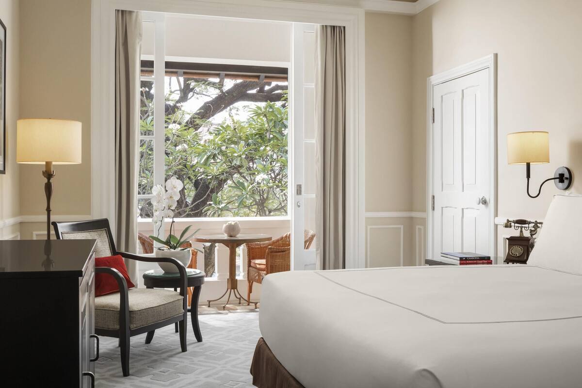 A hotel room with a bed, a desk, a chair, a lamp, a small table with a plant, and a view of greenery outside.