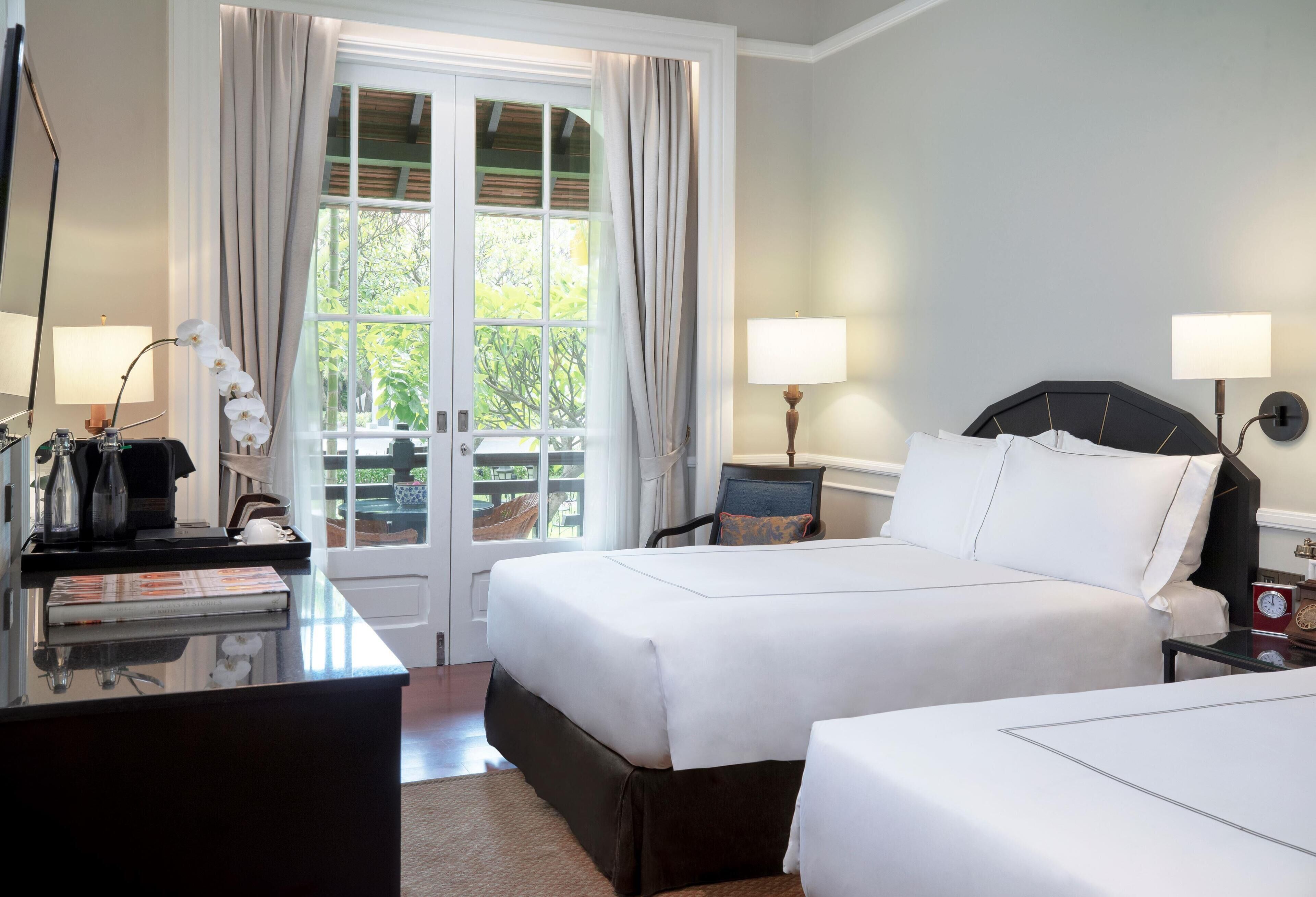 Une chambre d’hôtel avec deux lits, un bureau avec une lampe, une chaise et une vue sur la verdure par une fenêtre.