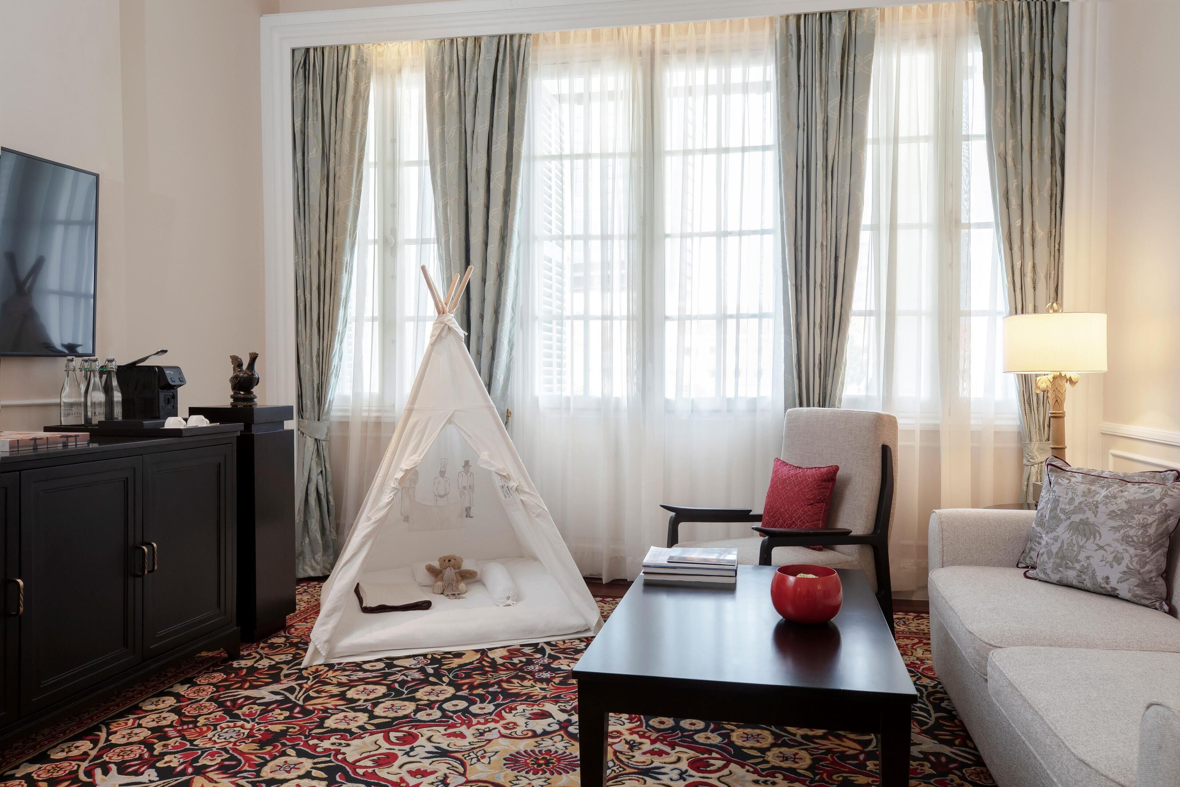 A living room with a sofa, a chair, a coffee table, a TV, a bookshelf, a tent, and a patterned carpet.