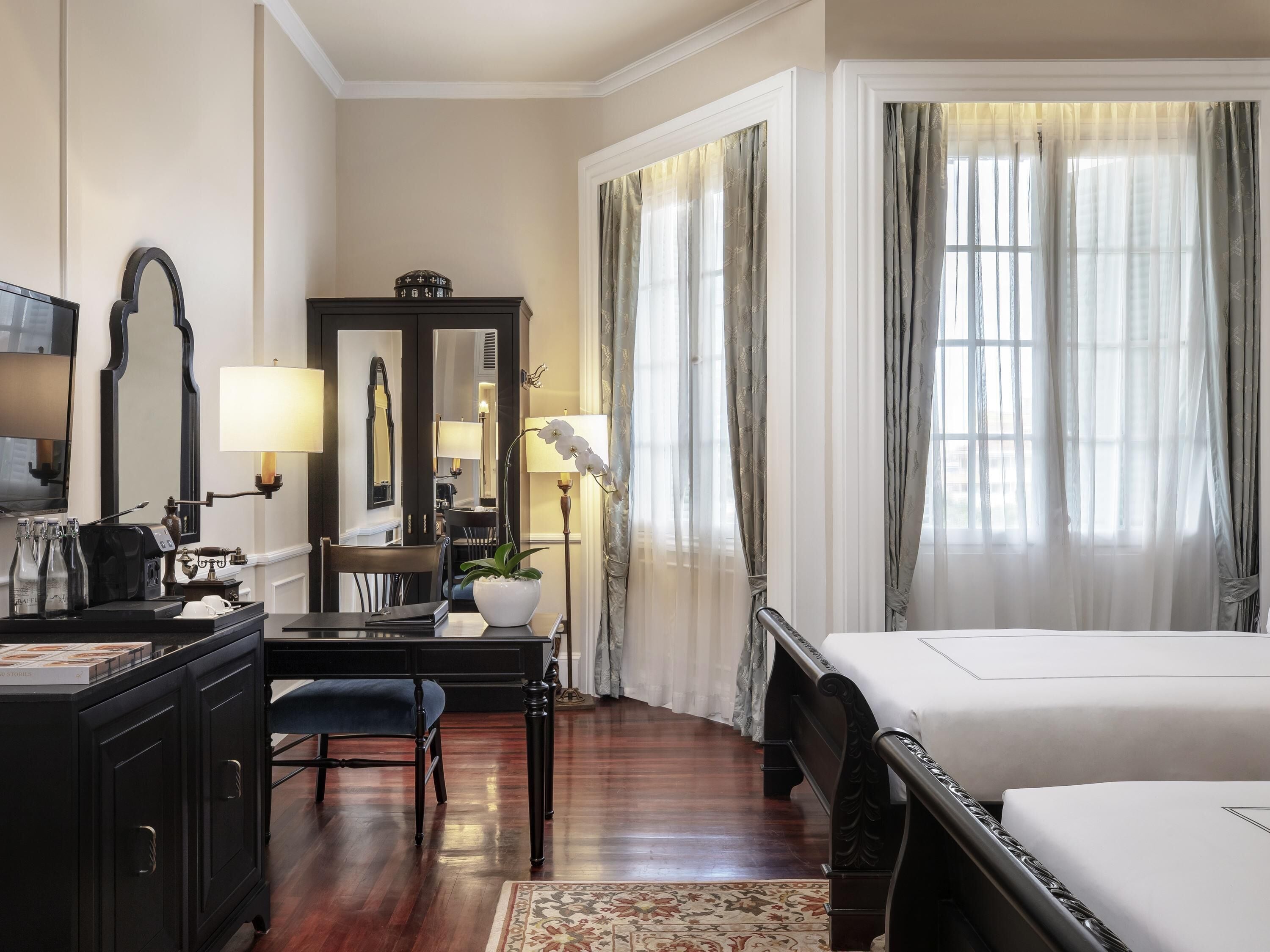 Habitación de hotel con cama, escritorio con lámpara, silla, cómoda, espejo y ventana con cortinas.