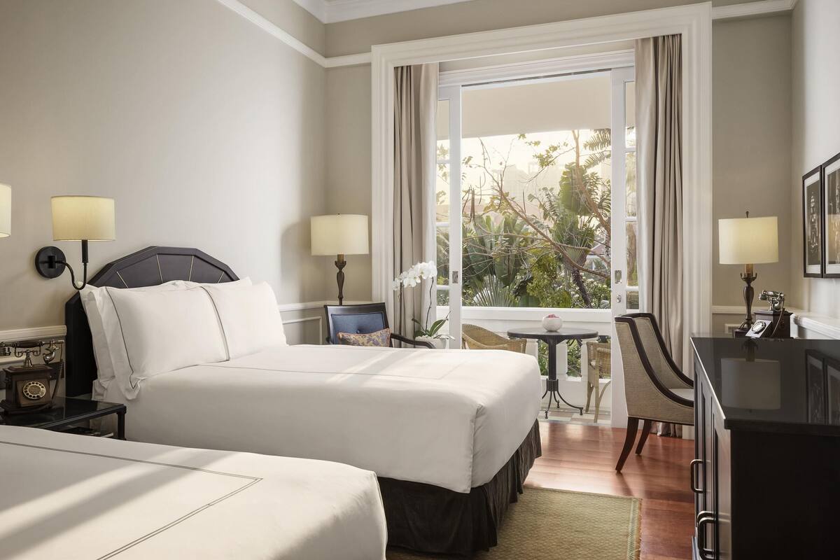 A hotel room with two beds, a desk, a chair, a small table, and a view of the outdoors.