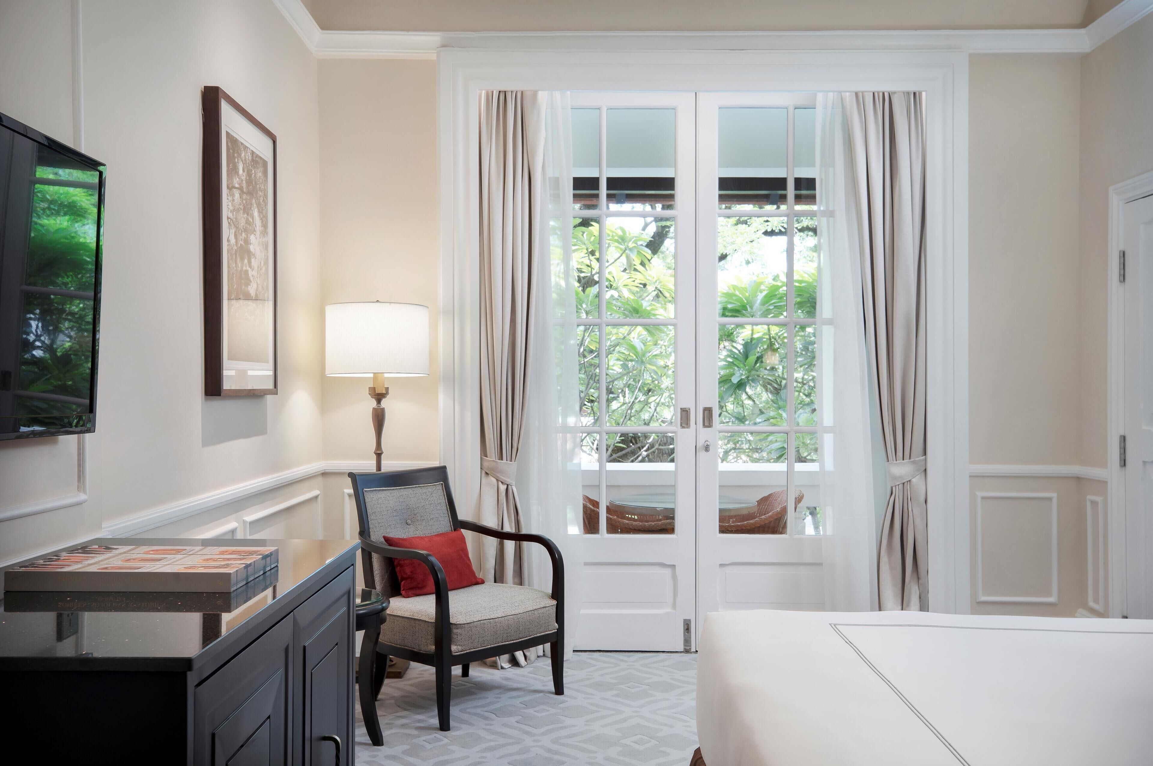 A hotel room with a large window, a desk with a lamp, a chair, and a bed.