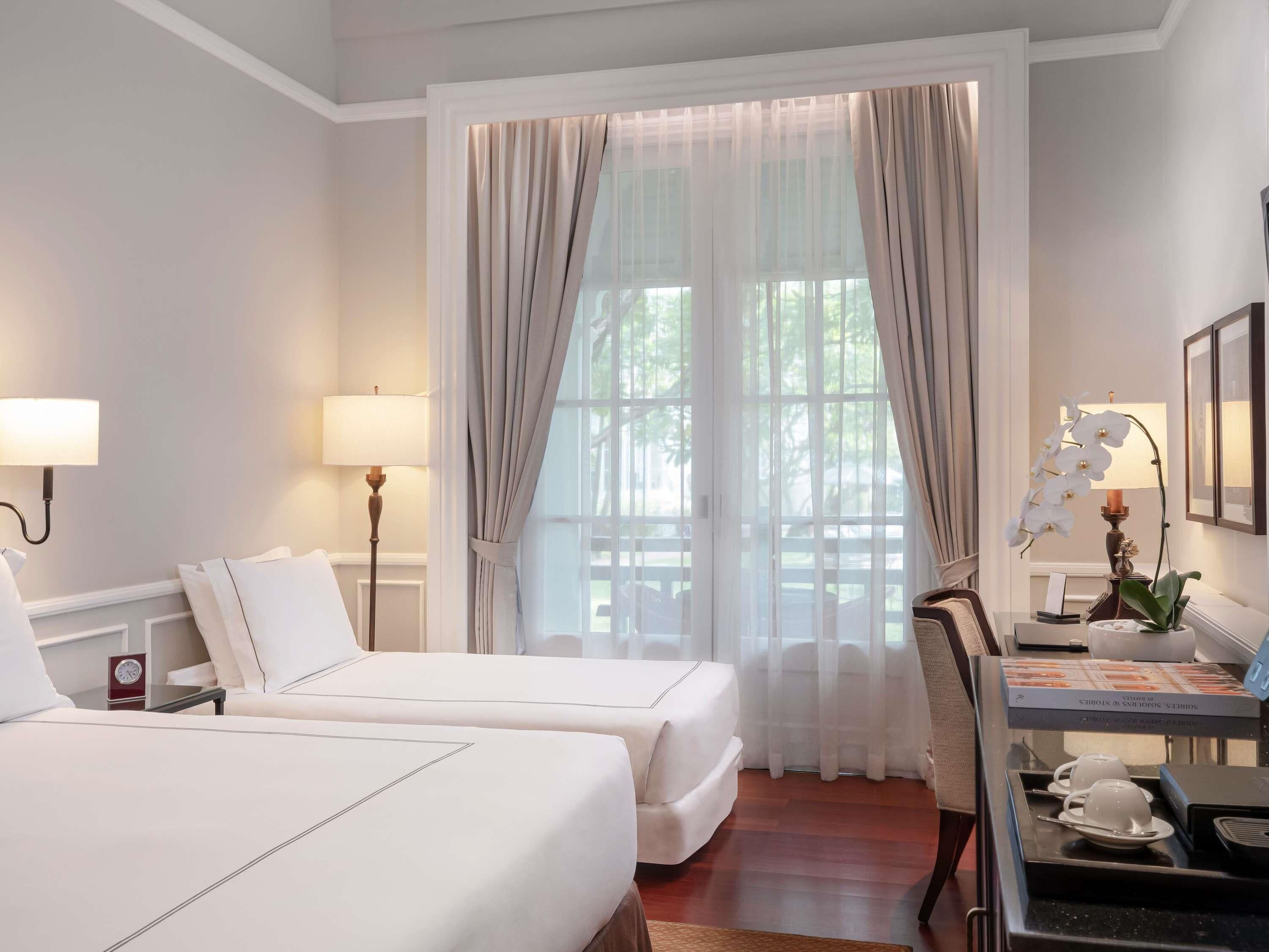 A hotel room with two beds, a desk with a lamp, a chair, and a view through large windows.