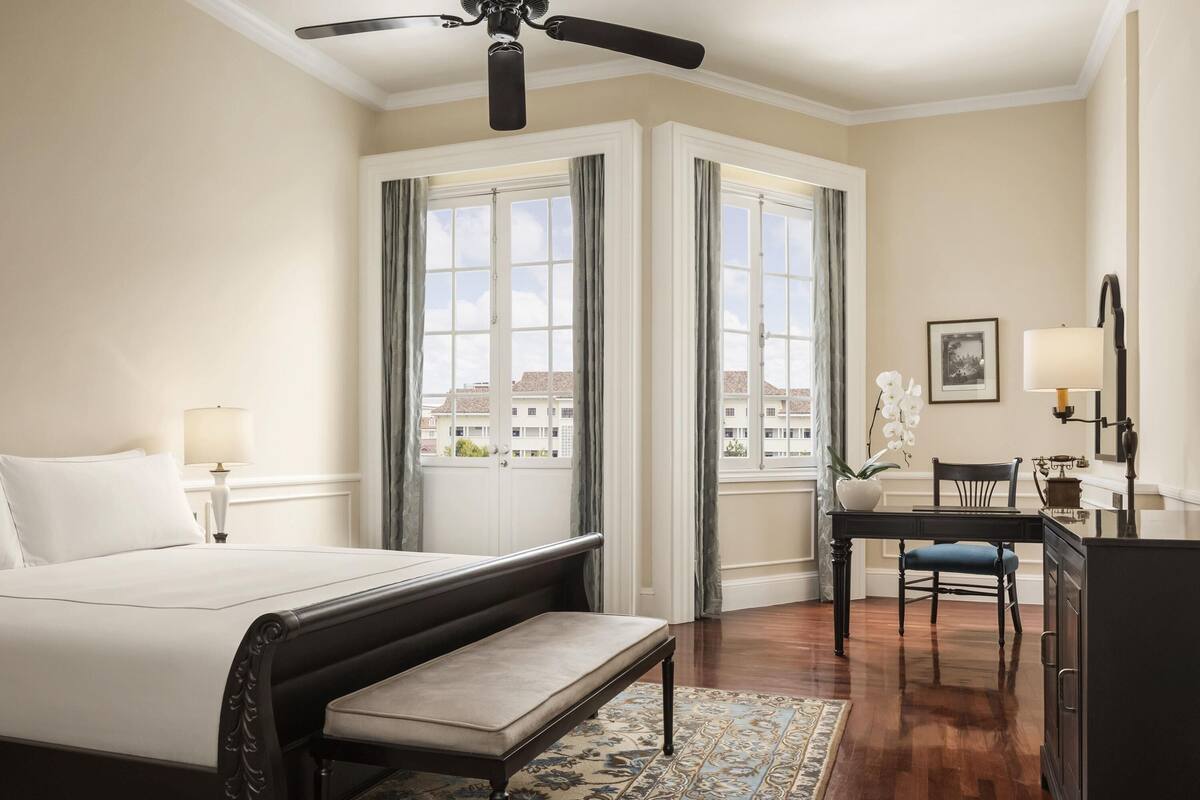 A hotel room with a bed, a desk, a chair, a bench, a ceiling fan, and large windows with curtains.