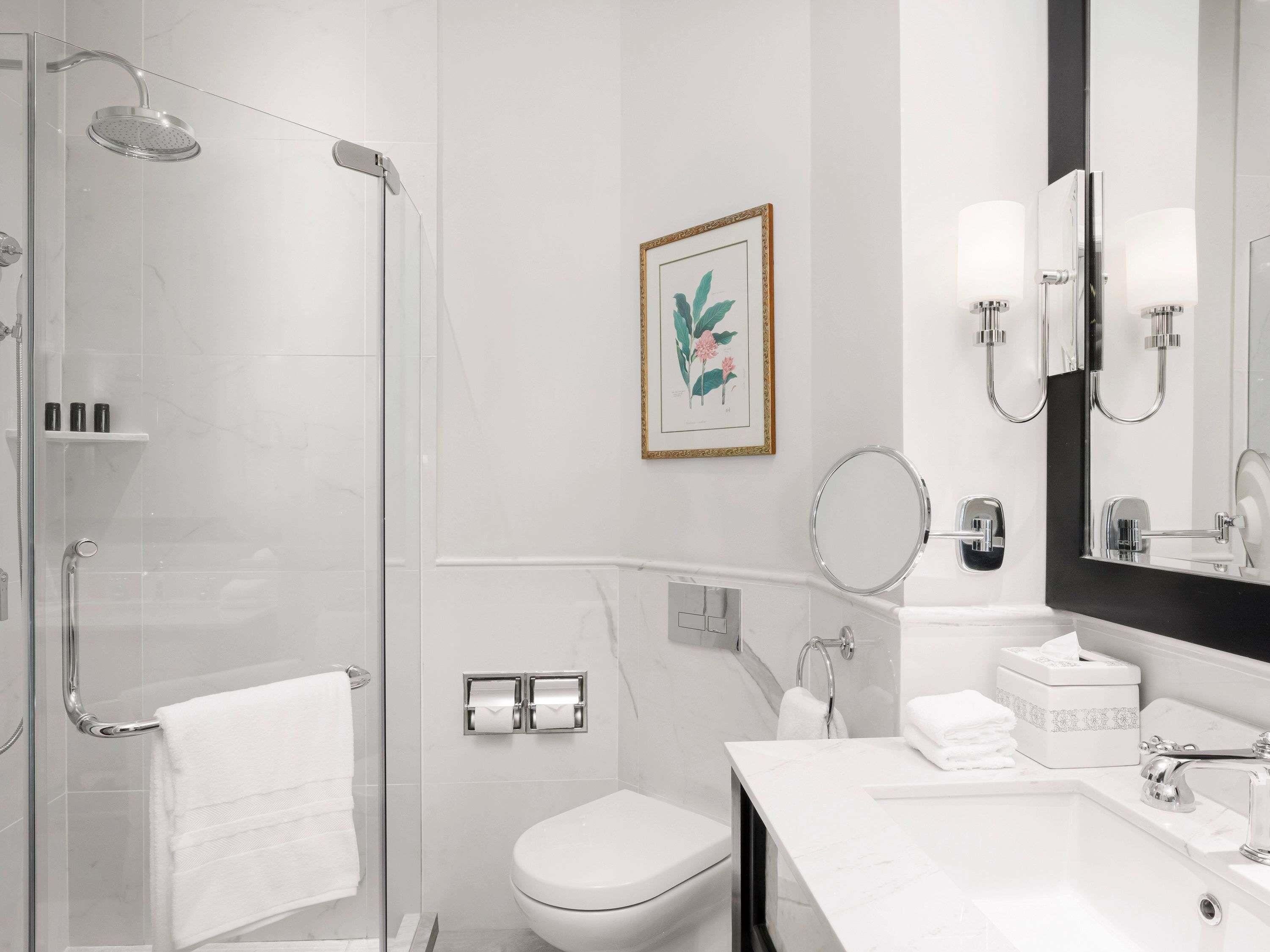 A modern bathroom with a glass shower enclosure, a toilet, a sink with a mirror, and a framed botanical print on the wall.