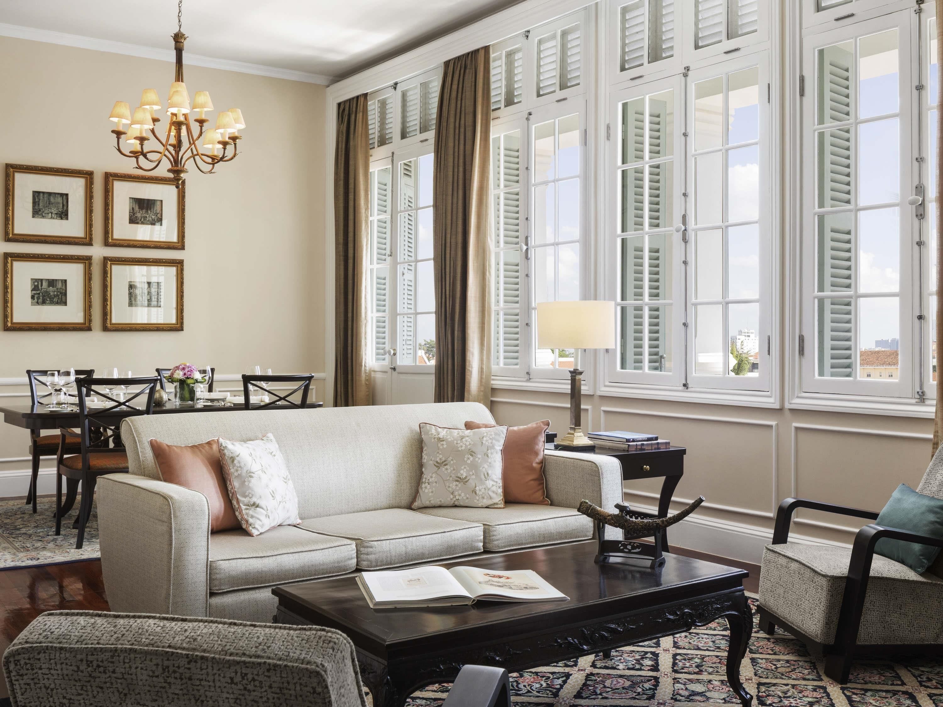 A living room with a sofa, coffee table, and armchairs, a dining area with a table and chairs, and large windows with shutters.