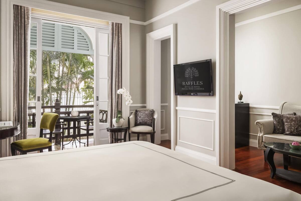 A hotel room with a large bed, a dining area with chairs and a table, a sofa, and a television displaying the hotel's name.