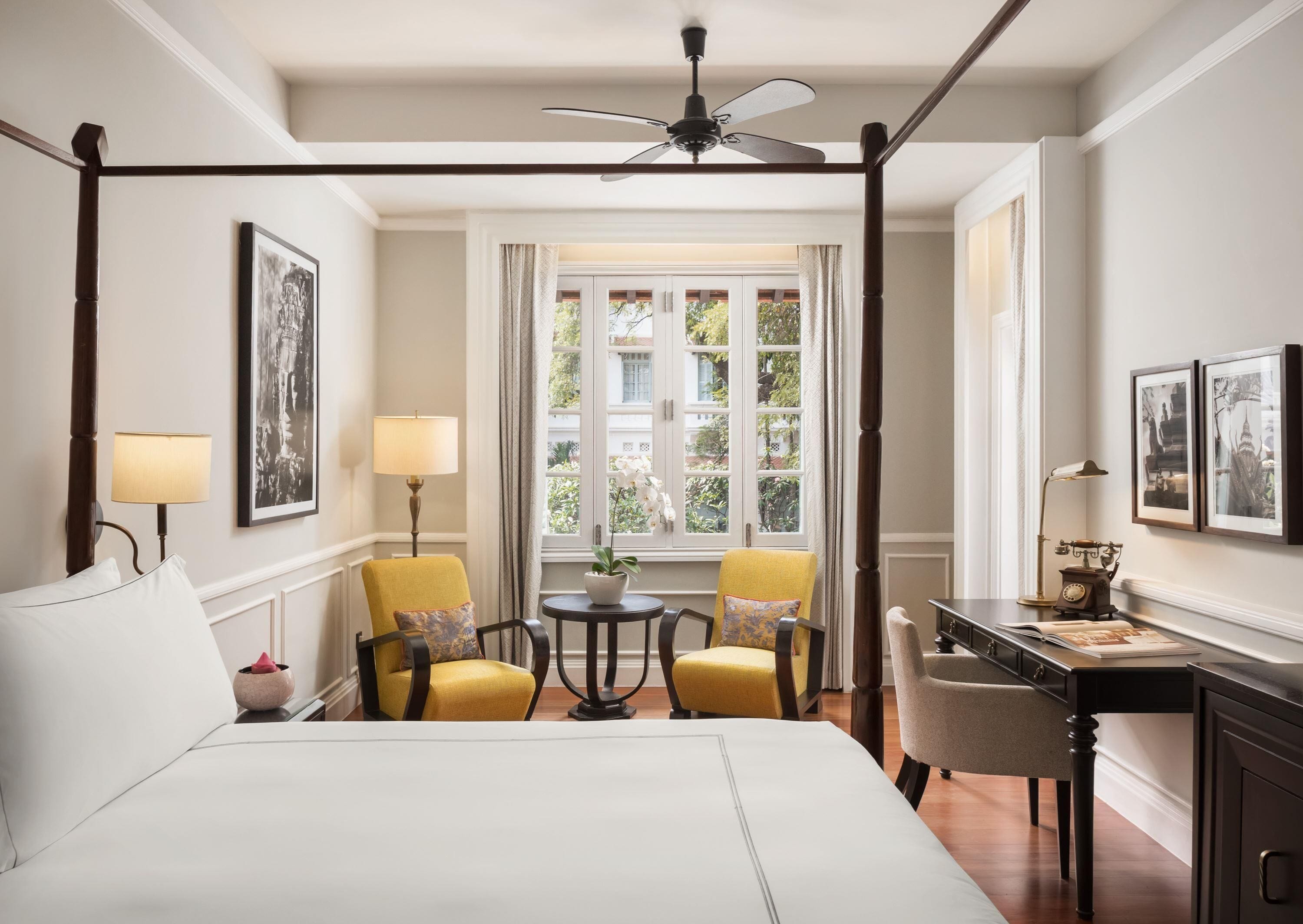 A hotel room with a four-poster bed, two chairs, a desk, a ceiling fan, and a window with curtains.