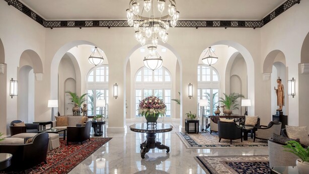 Eine großzügige Lobby mit hohen Decken, Rundbögen und einem zentralen Kronleuchter. Es gibt Sitzbereiche mit Sofas und Sesseln, einen runden Tisch mit Blumenvase und eine Statue auf Sockel.
