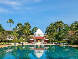 A pool with lounge chairs and umbrellas, surrounded by palm trees and a white building with a red roof.