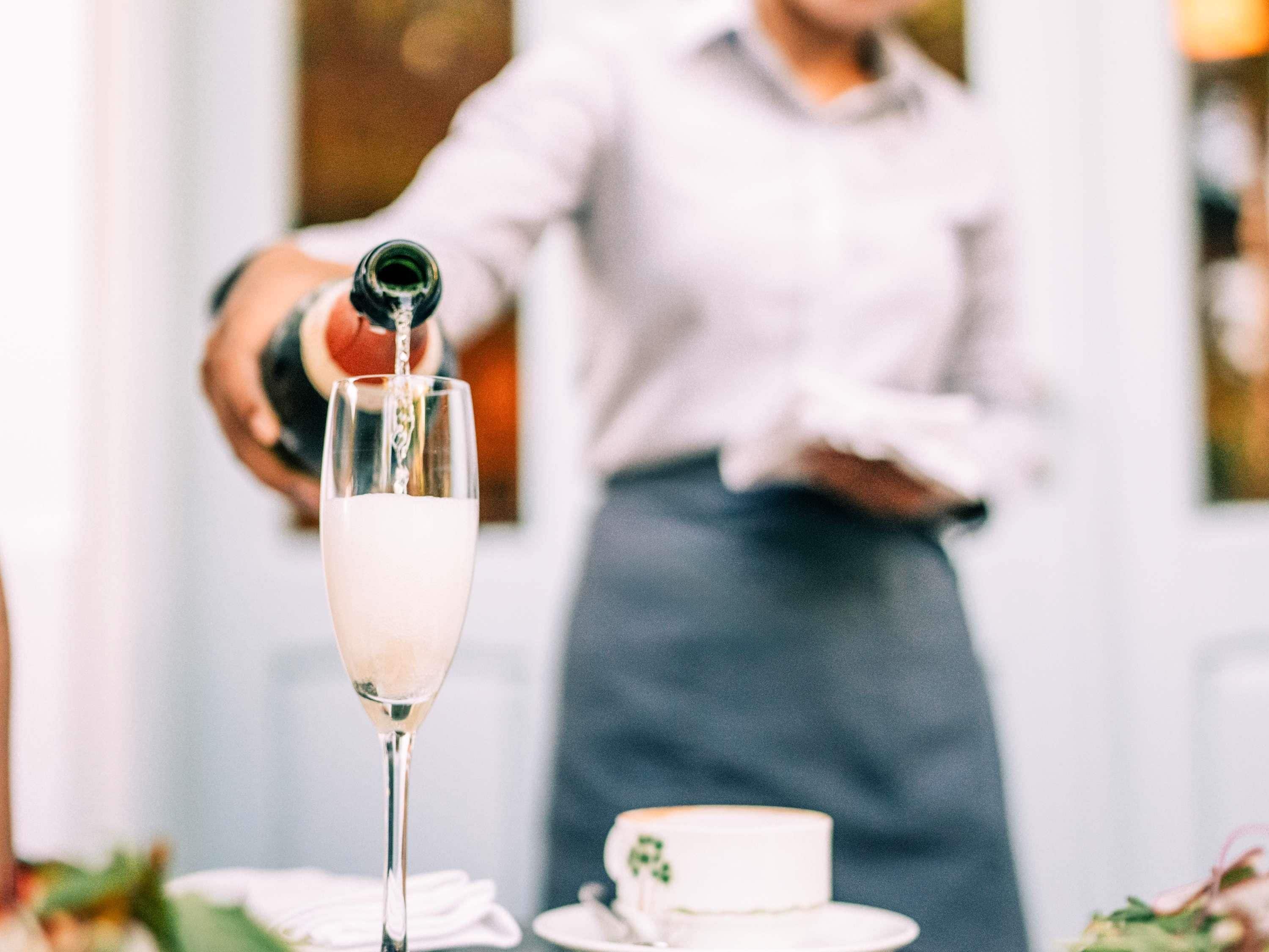 Un verre de champagne en cours de remplissage, un gâteau sur une assiette et une personne à l’arrière-plan.