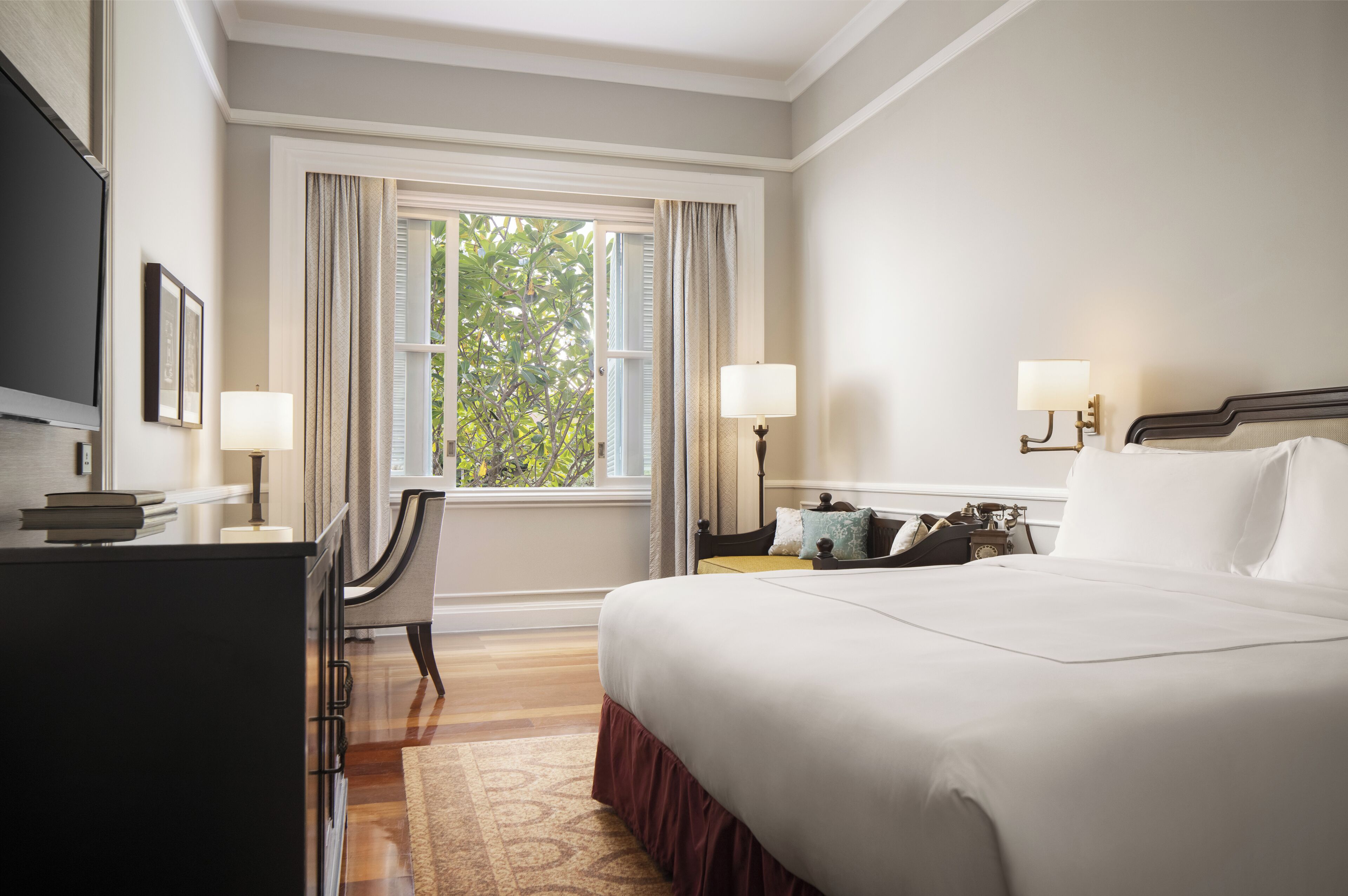 A hotel room with a large bed, two bedside tables with lamps, a desk with a chair, and a window with curtains.