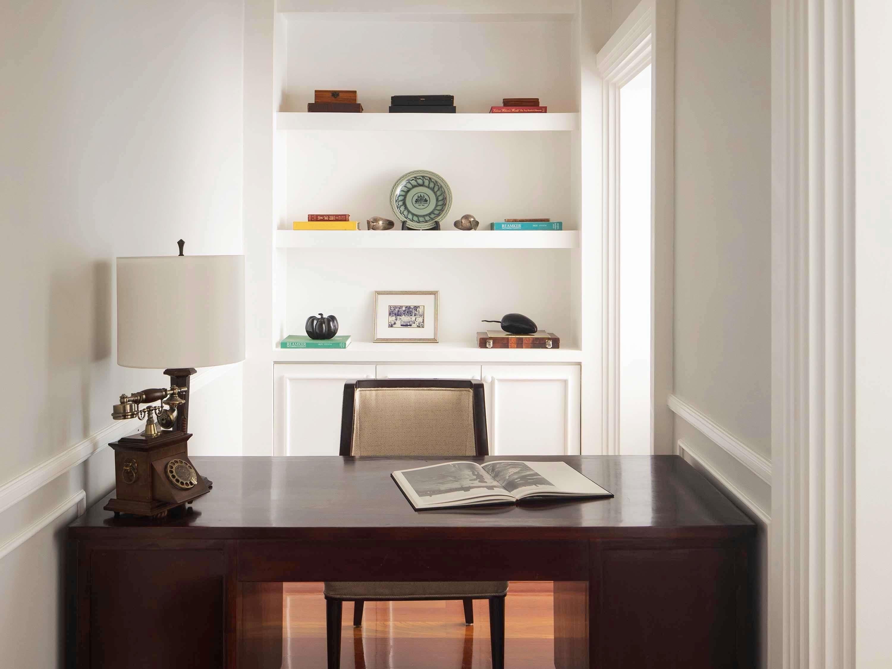 A wooden desk with a lamp, a chair, and a book. There is a fireplace with a mantel and shelves above it.