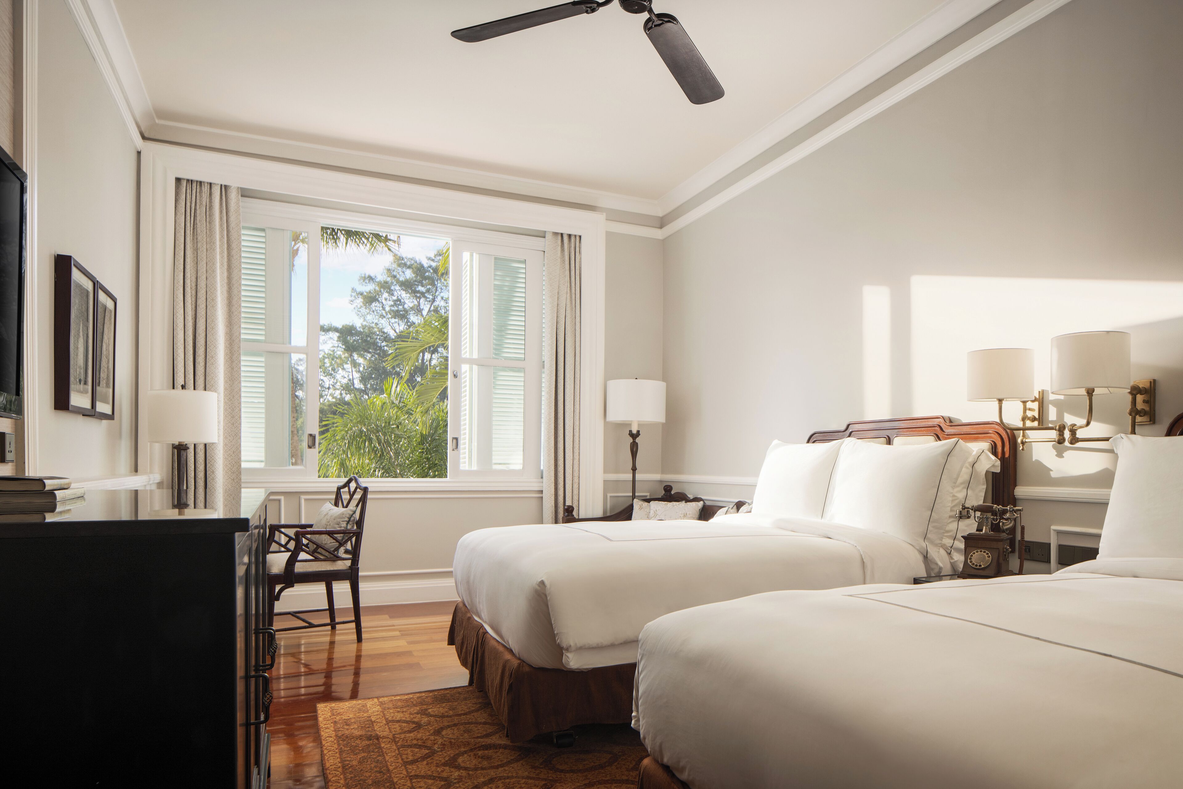 A hotel room with two beds, a desk, a chair, a ceiling fan, and a window with shutters.