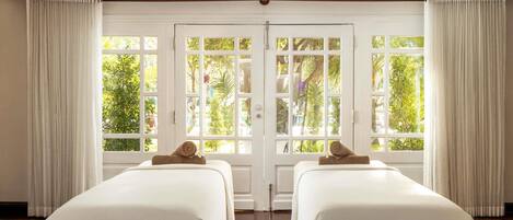 A room with two beds, a large window with curtains, and a view of greenery outside.