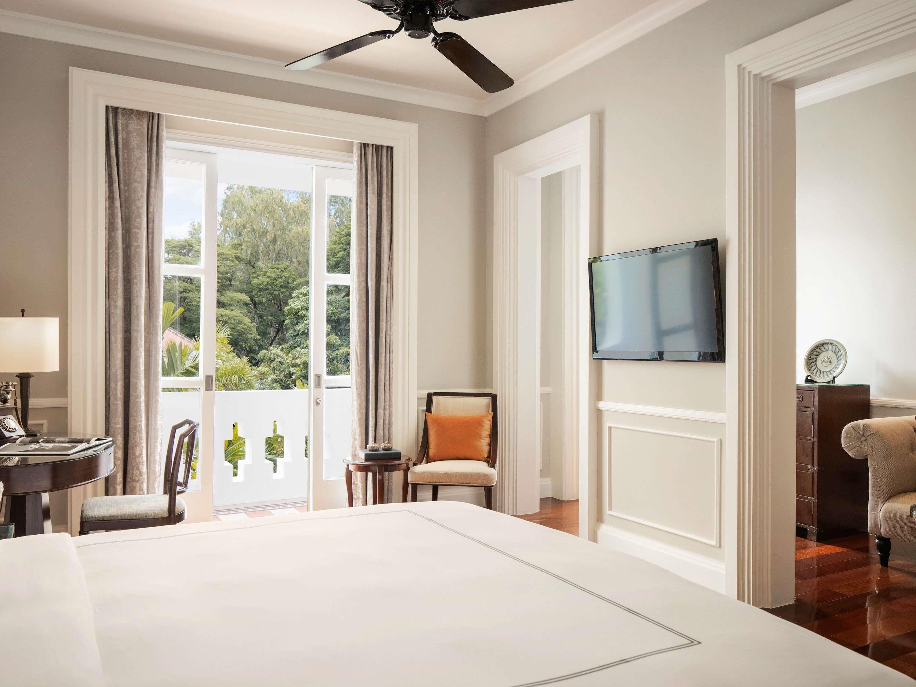 A hotel room with a large bed, a television mounted on the wall, a desk with a lamp, and a view of greenery outside.